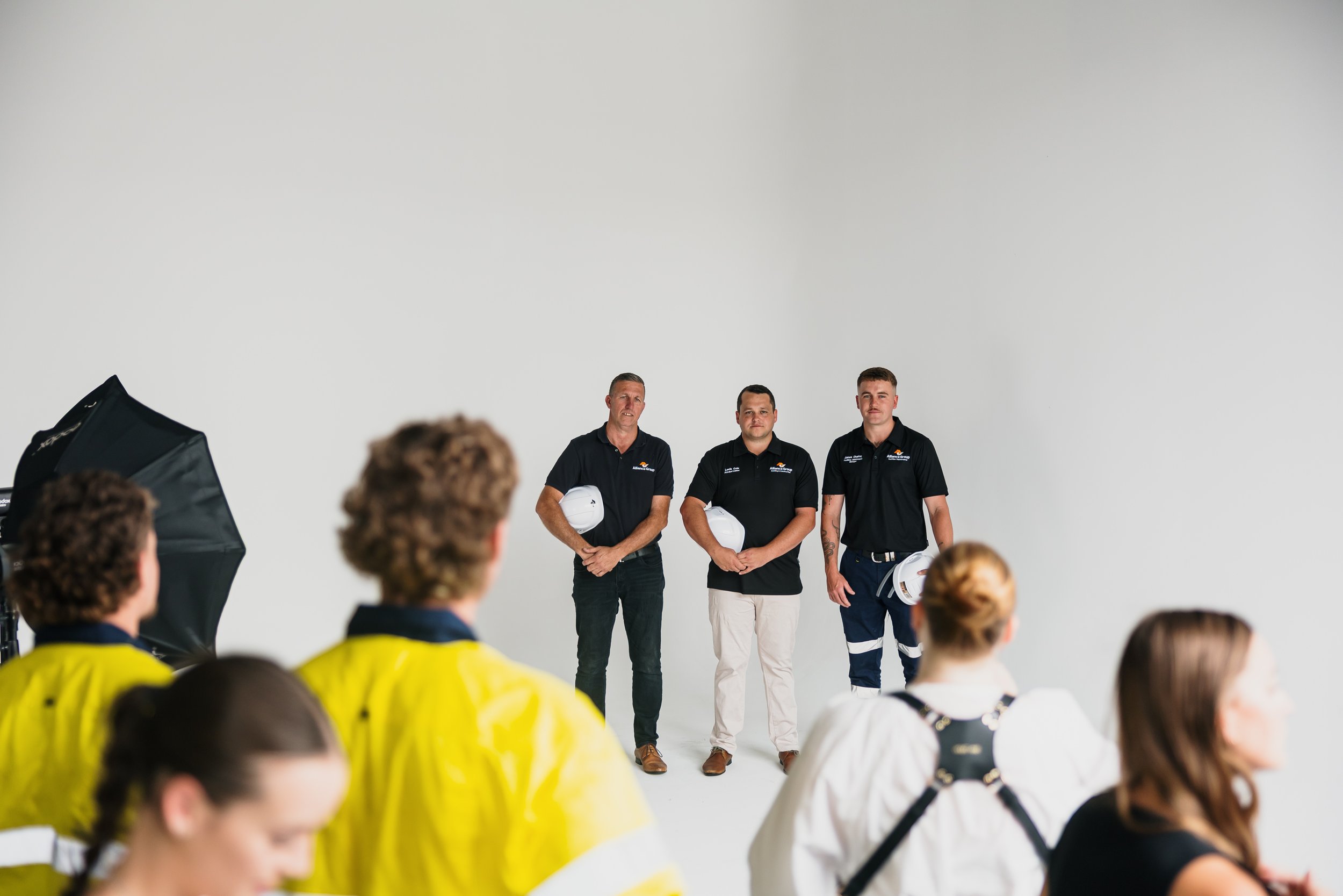 Three men in black shirts holding white helmets stand against a plain white background, with a diverse audience in safety gear seated in front of them during a presentation or event.