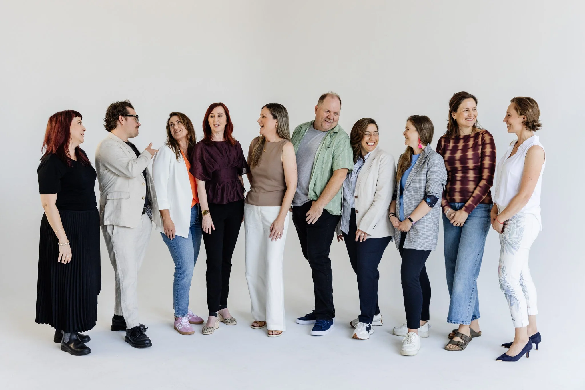 Group of ten diverse people standing in a line, smiling, engaging in conversation, against a plain white background.