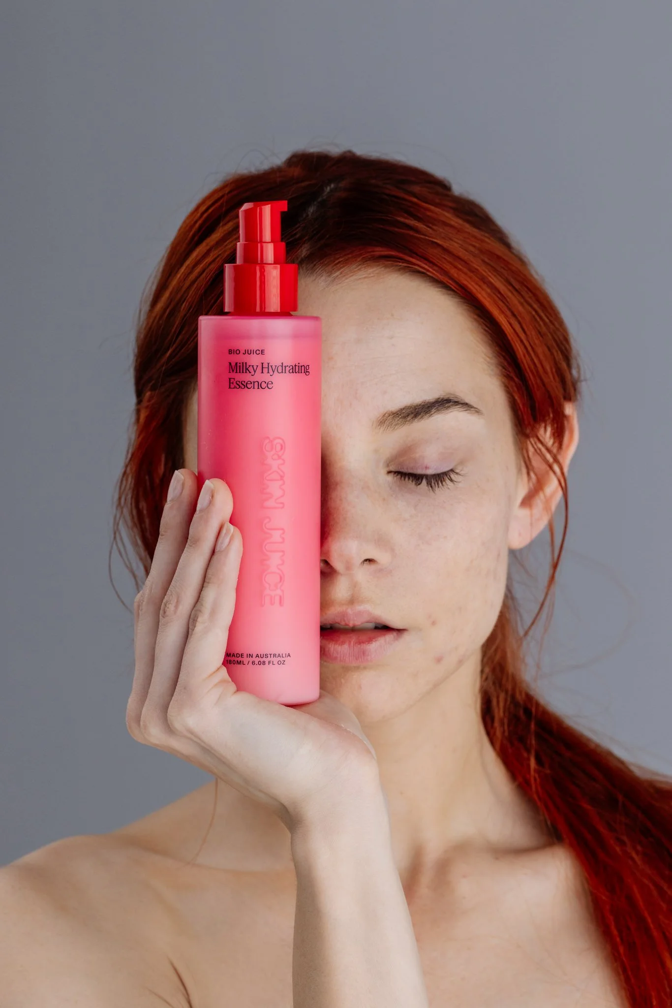A woman with red hair holding a pink bottle of skincare product in front of her face with her eyes closed.