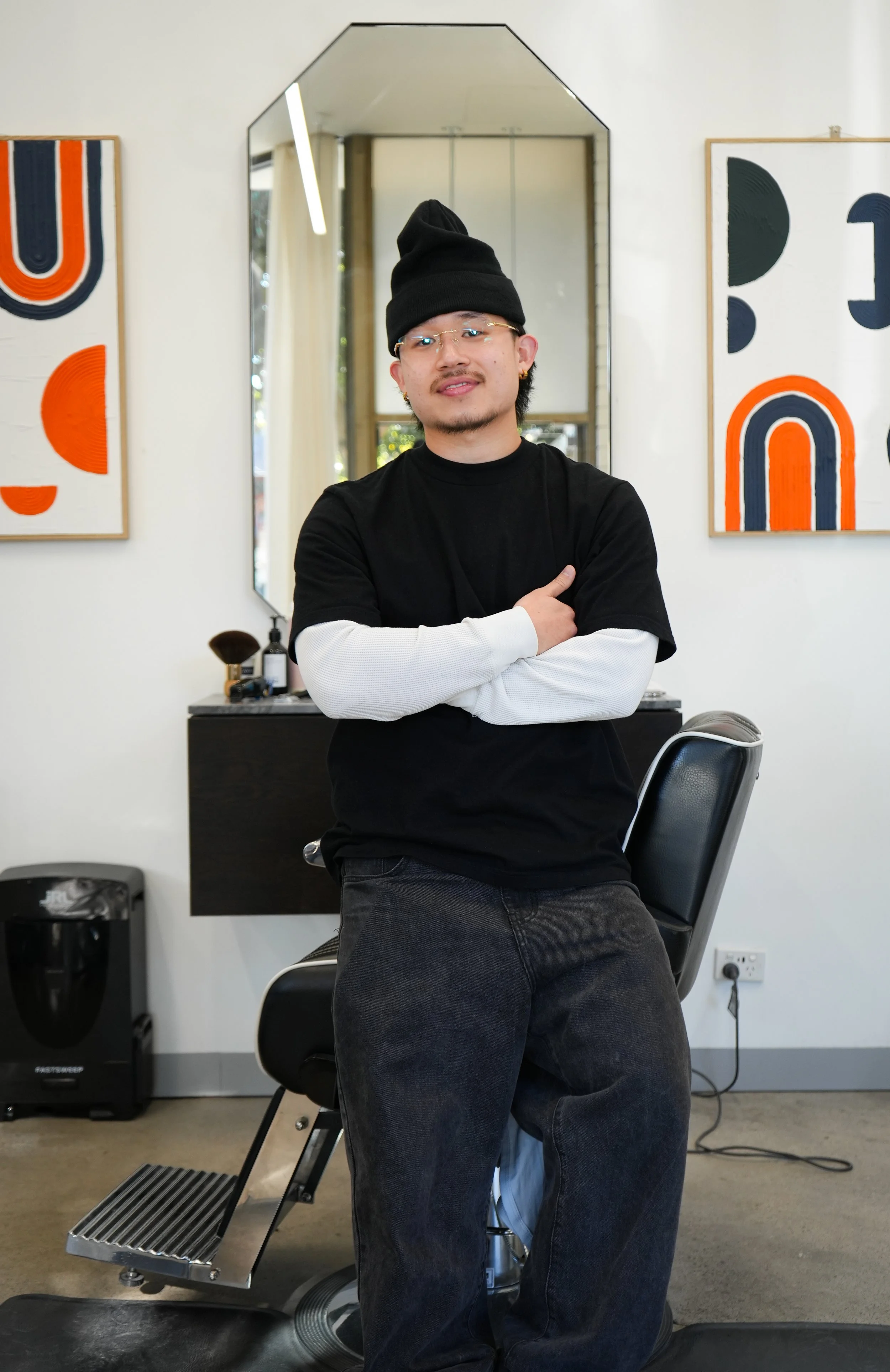 A man wearing a black beanie, glasses, and layered black and white clothing, sitting on a black chair in a salon or barber shop, with art on the walls, a mirror, and hair products in the background.