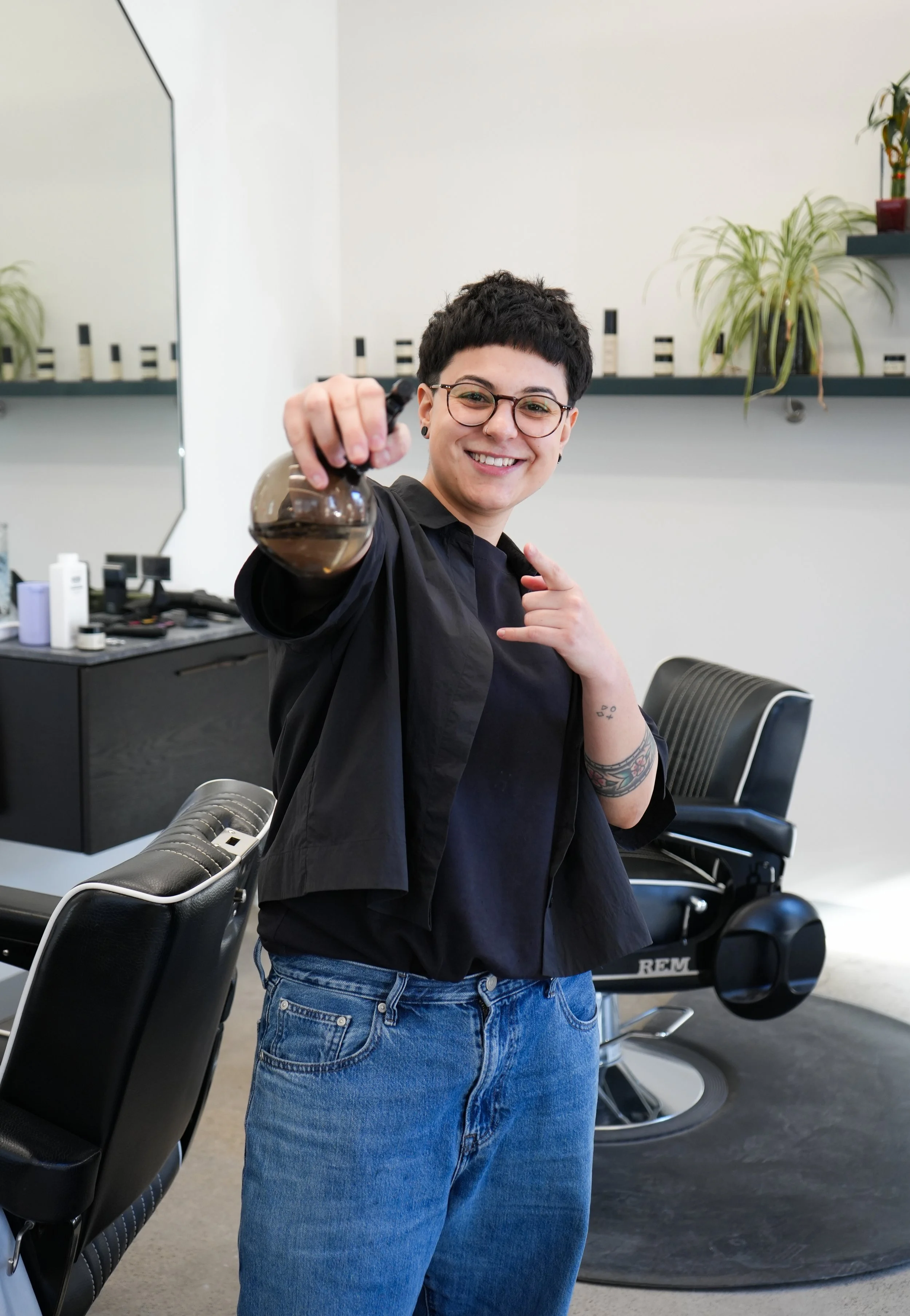 A woman with short dark hair and glasses smiling, holding a coffee pot in a hair salon or barbershop with chairs, mirror, and plants in the background.