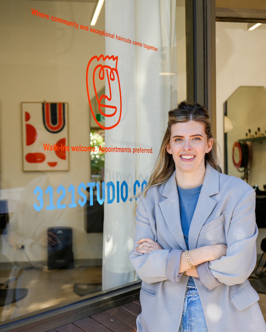 A young woman standing outside a studio's glass door, smiling with arms crossed, the door has colorful text and illustrations indicating walk-in appointments are welcome, and the studio's website is 3121STUDIO.COM.