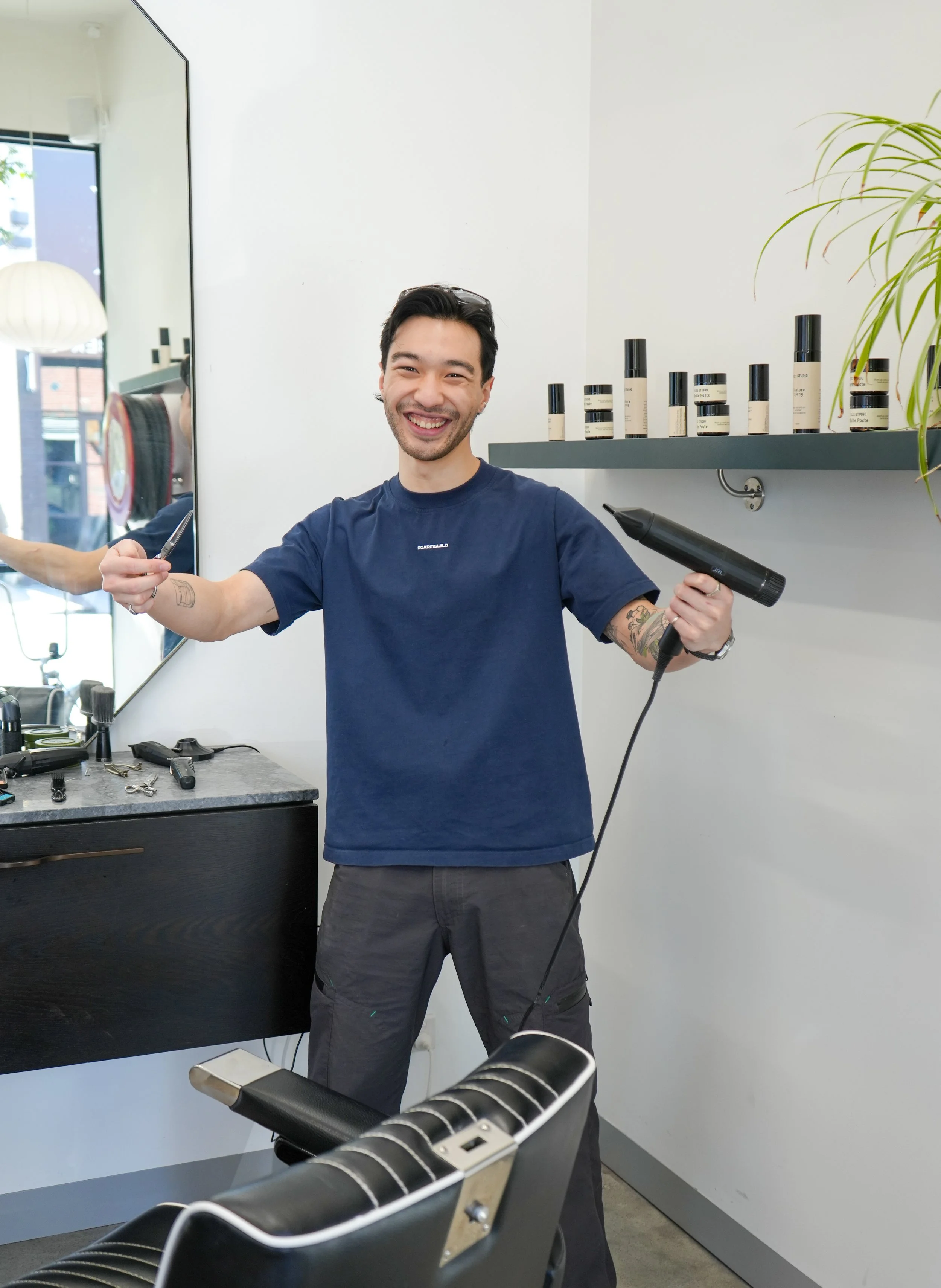 A man is smiling and holding a hairdryer and a hairbrush in a barbershop or salon. There are bottles of hair products on a shelf behind him and a mirror reflecting part of the room.