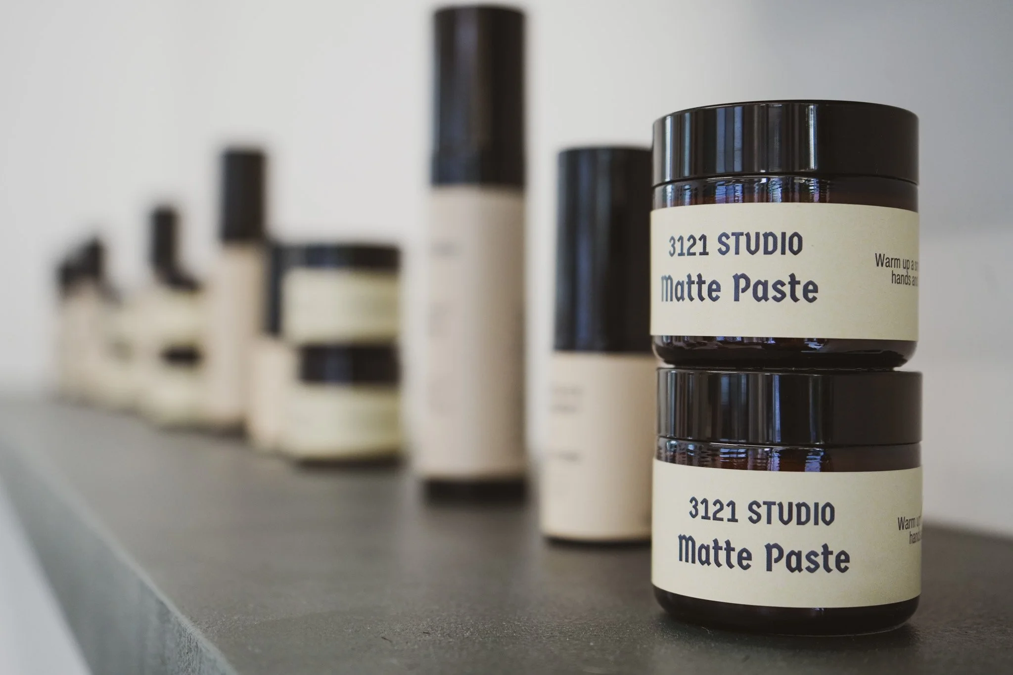 Close-up of a jar labeled '3121 Studio Matte Paste' on a gray shelf, with other blurred hair styling products in the background.