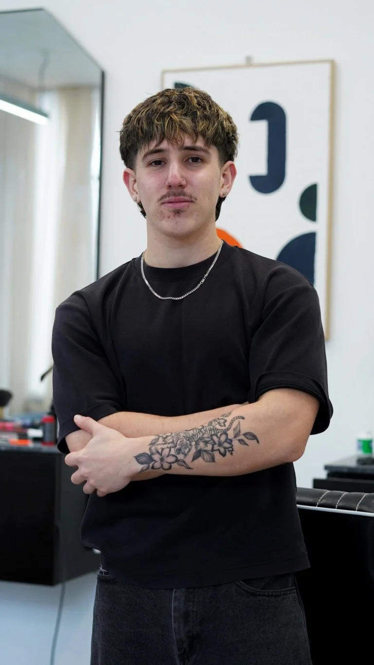 A young man with short, curly hair and a mustache, wearing a black T-shirt and a silver chain, stands with arms crossed, showing a tattoo of a tiger and flowers on his left forearm, in a modern indoor setting.