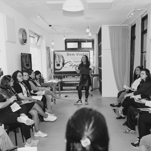 Mulheres sentadas em círculo participam de um workshop em uma sala iluminada, enquanto uma facilitadora, em pé ao centro, fala e gesticula. Ao fundo, uma faixa com os dizeres “Bem-Vindas” está pendurada.