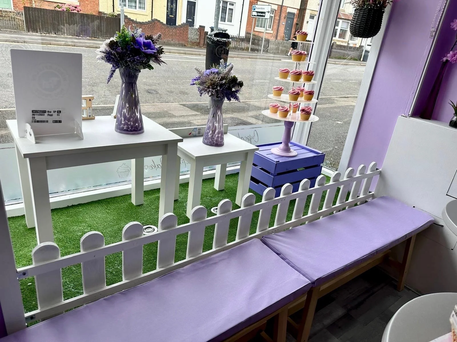 Interior of a cafe decorated with purple flowers, pink cupcakes on a stand, and purple benches, with a large window showing a street outside.