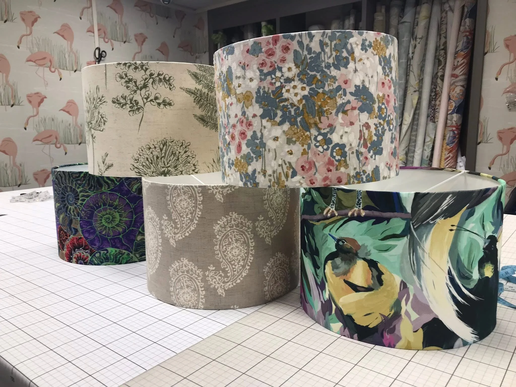 Six fabric lampshades with various floral and nature-themed patterns on a grid-lined table. In the background, there are curtains and wallpaper with flamingo illustrations.