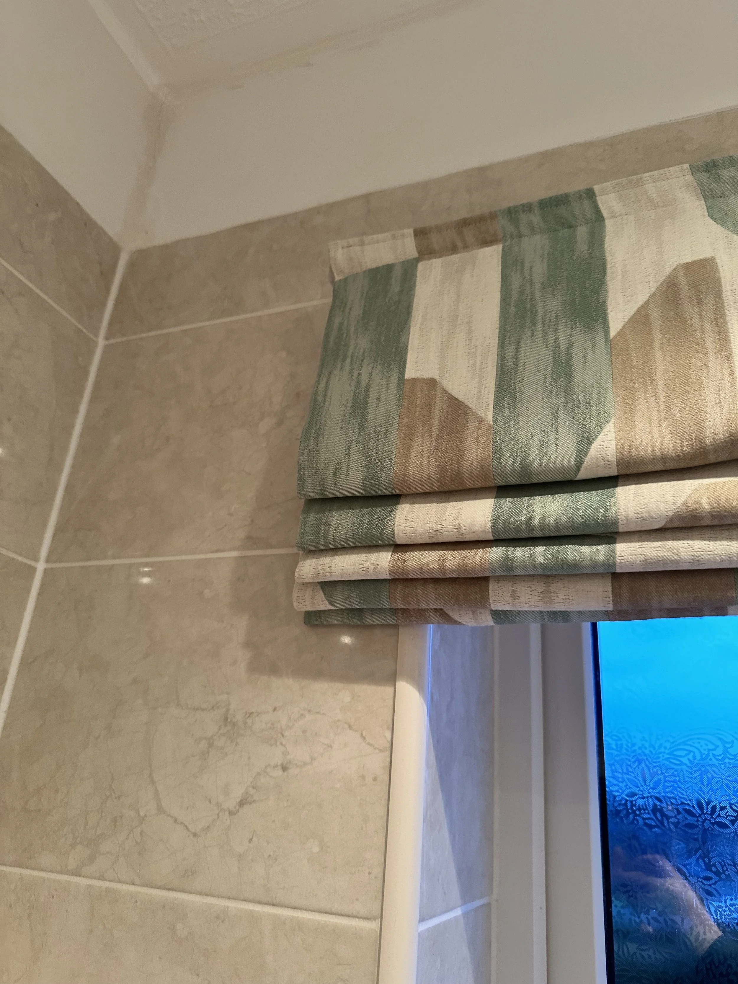 A window with a colorful striped roman shade in beige, green, and brown in a bathroom with beige tiled walls.