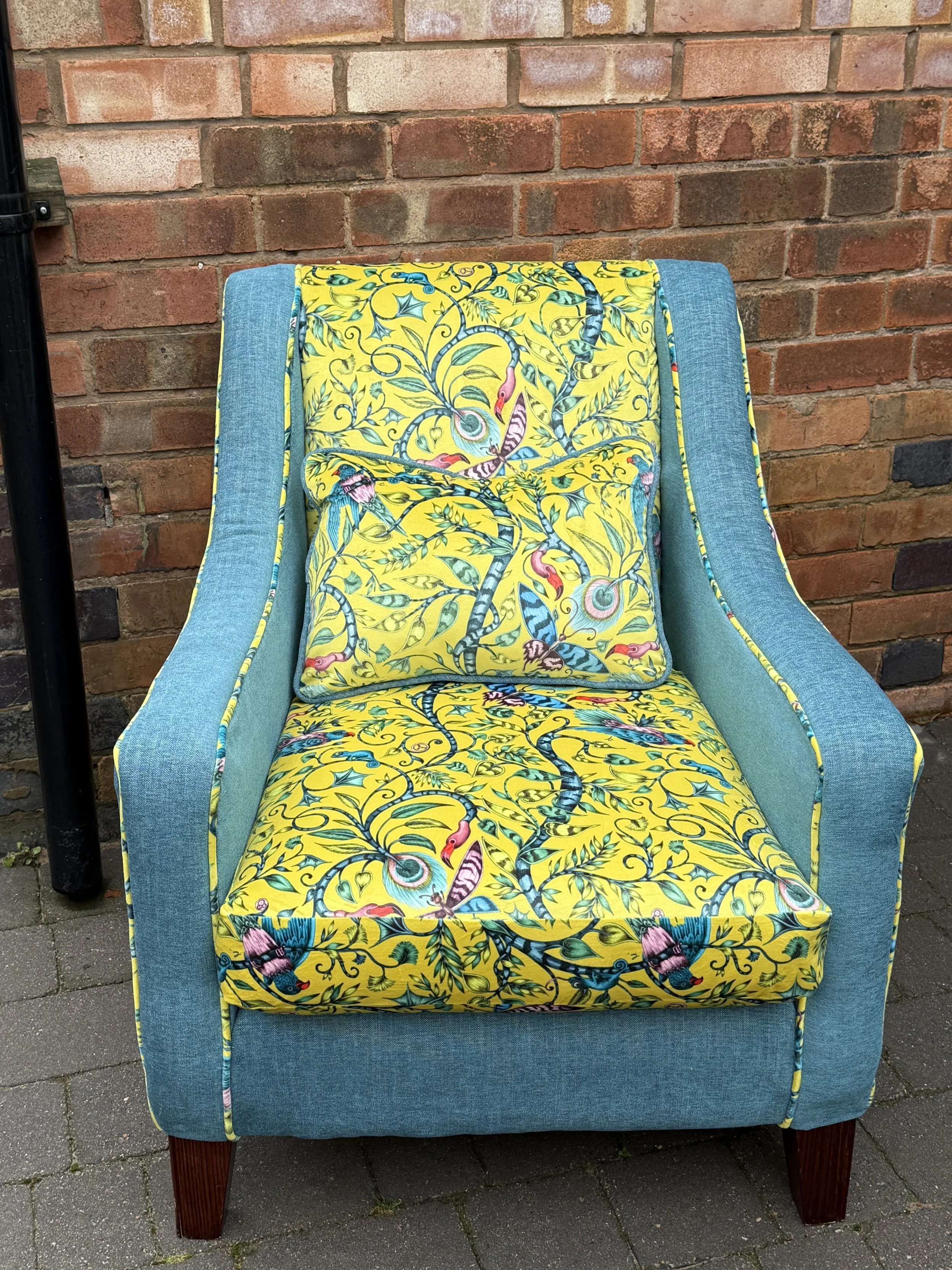 A wooden armchair with teal fabric on the sides and a colorful, yellow fabric with tropical designs on the seat and pillow.