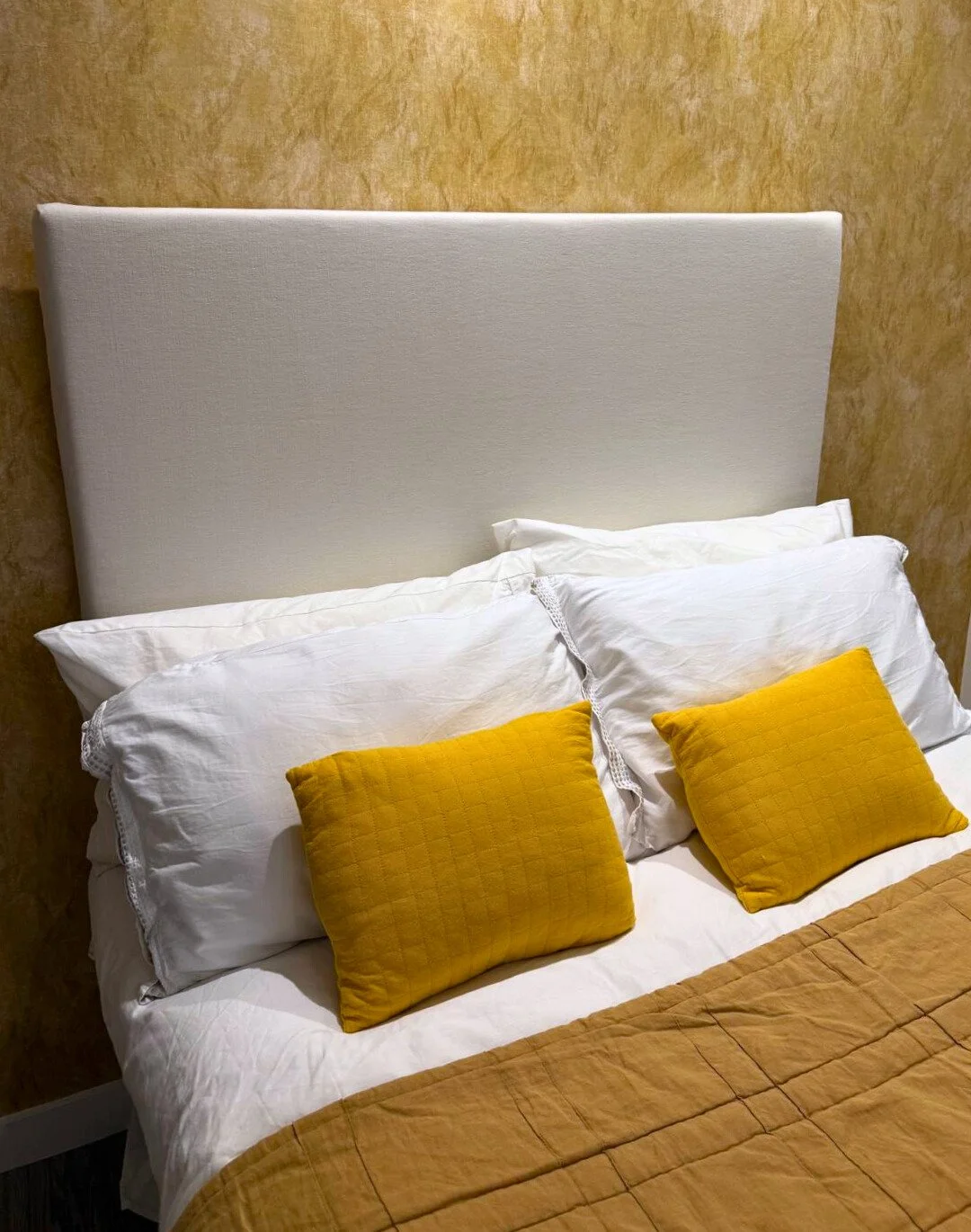 A bed with white sheets, two white pillows, two yellow throw pillows, a beige bedspread, and a white upholstered headboard against a textured beige wall.