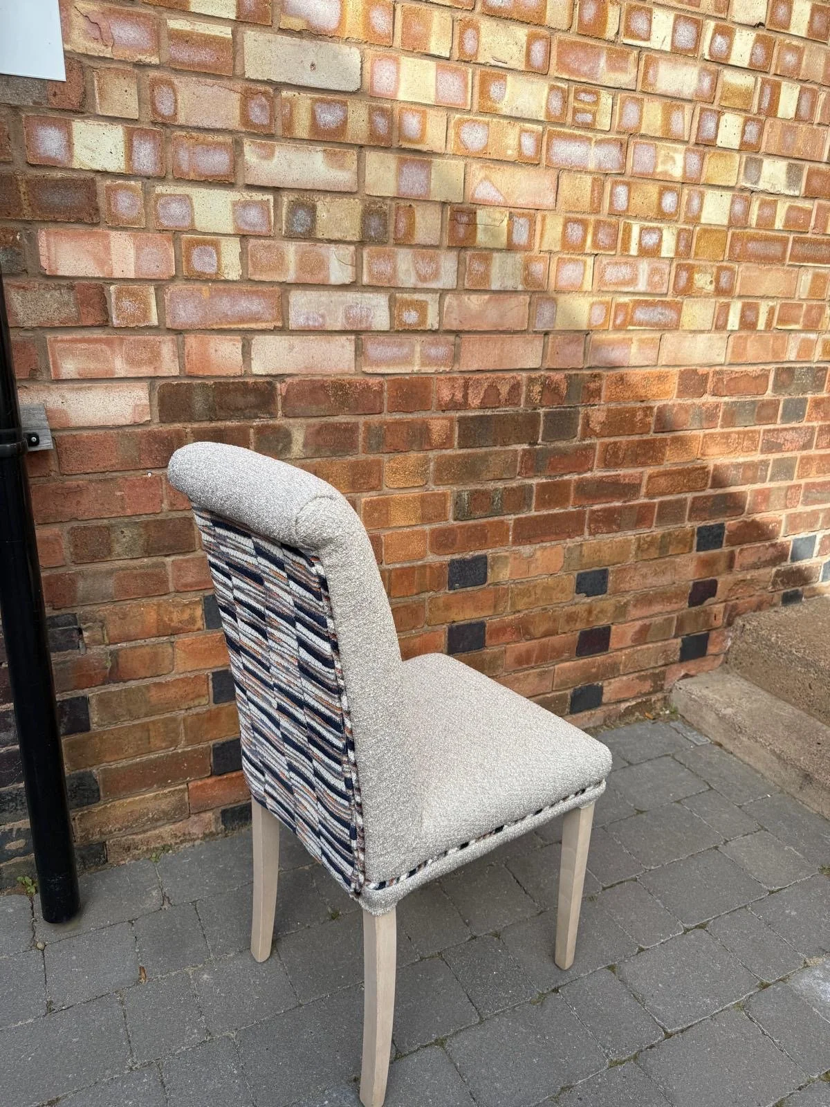 A chair with a cushioned seat and backrest, covered in a patterned fabric, is placed against a brick wall on a paved outdoor area.