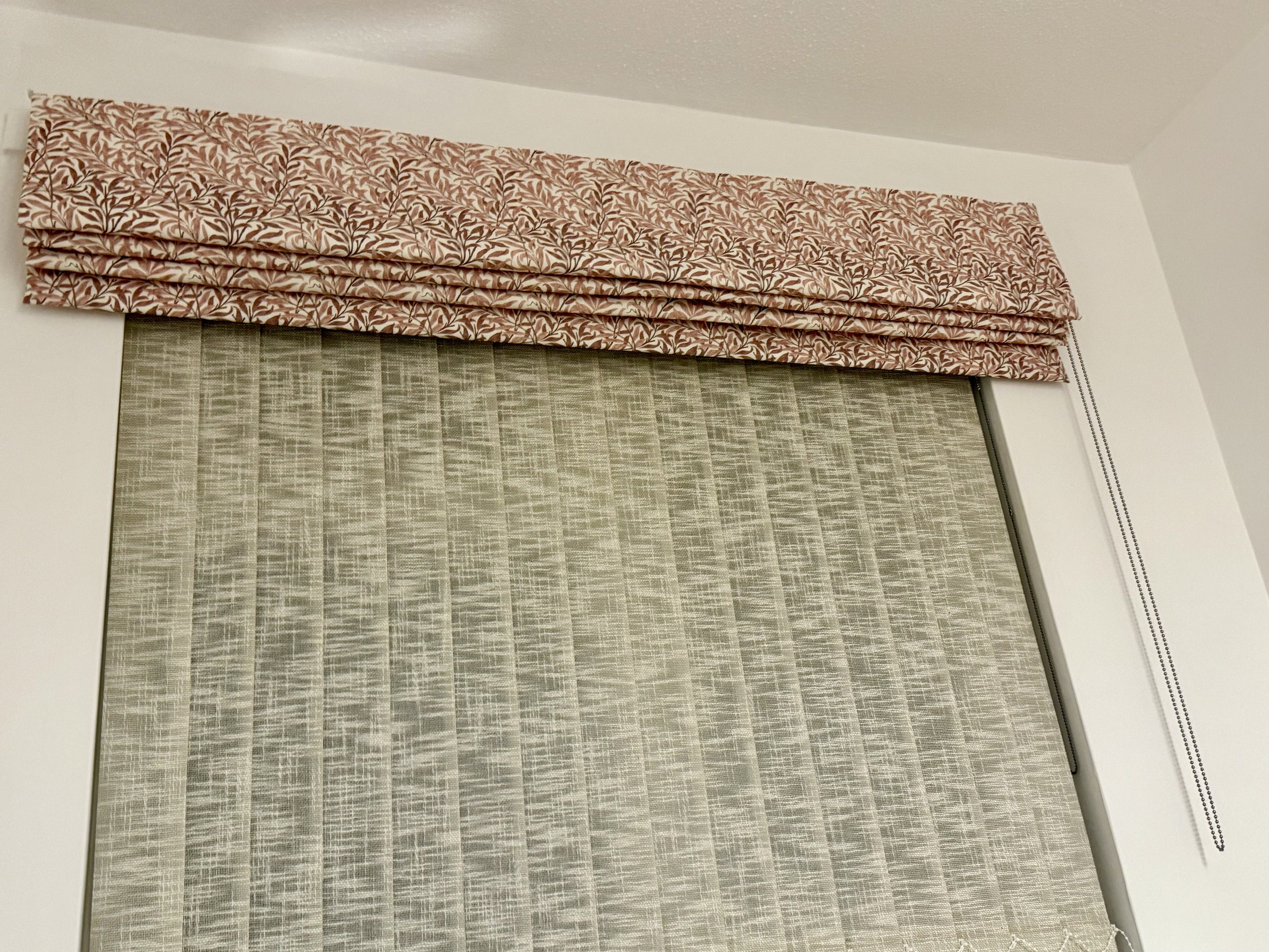 Window with beige patterned blinds and a pink floral valance at the top.