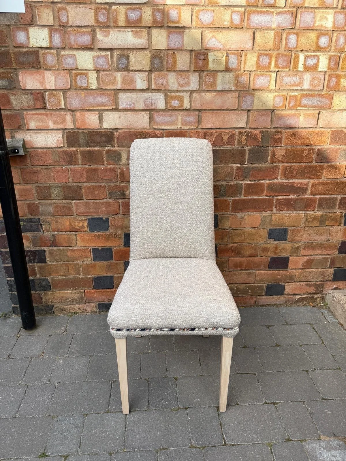 A beige upholstered chair with four light wooden legs, placed on gray brick pavement against a red brick wall.
