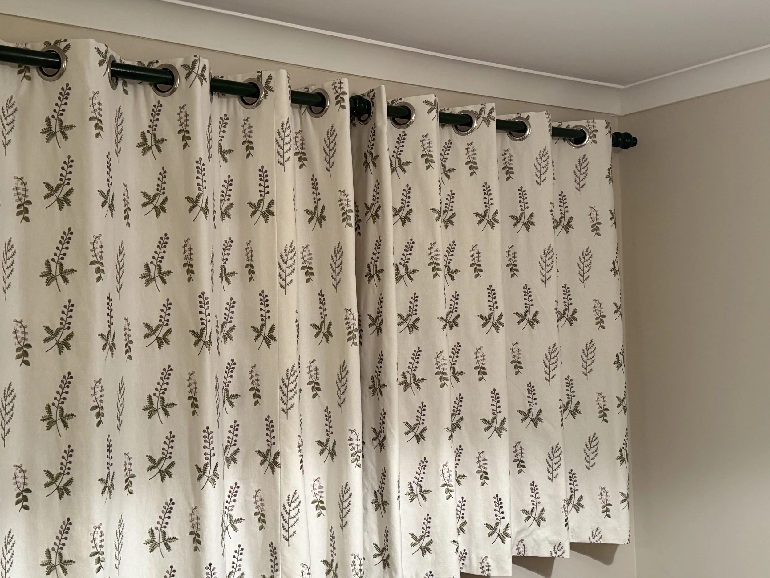 Cream-colored curtains with a floral pattern of purple and green leaves hanging on a dark rod in a room with a white ceiling and beige wall.