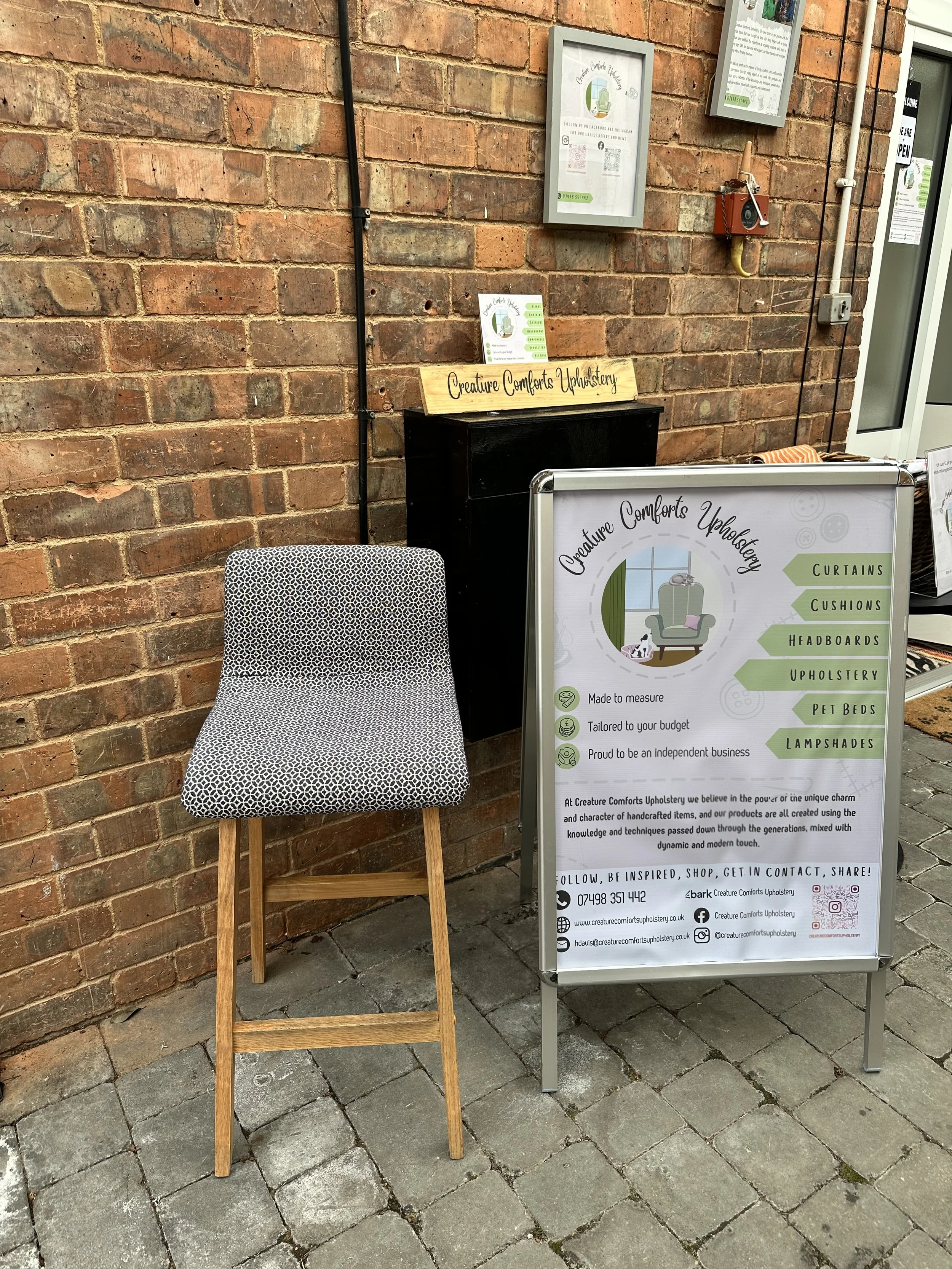 A sidewalk display for Creature Comforts Upholstery with a chair and an informational signboard listing services such as curtains, cushions, headboards, upholstery, pet beds, and lampshades, set against a brick wall.