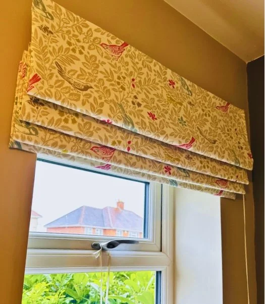 Close-up of a window with a floral and bird-patterned Roman shade partially lowered, showing a roof and some greenery outside.
