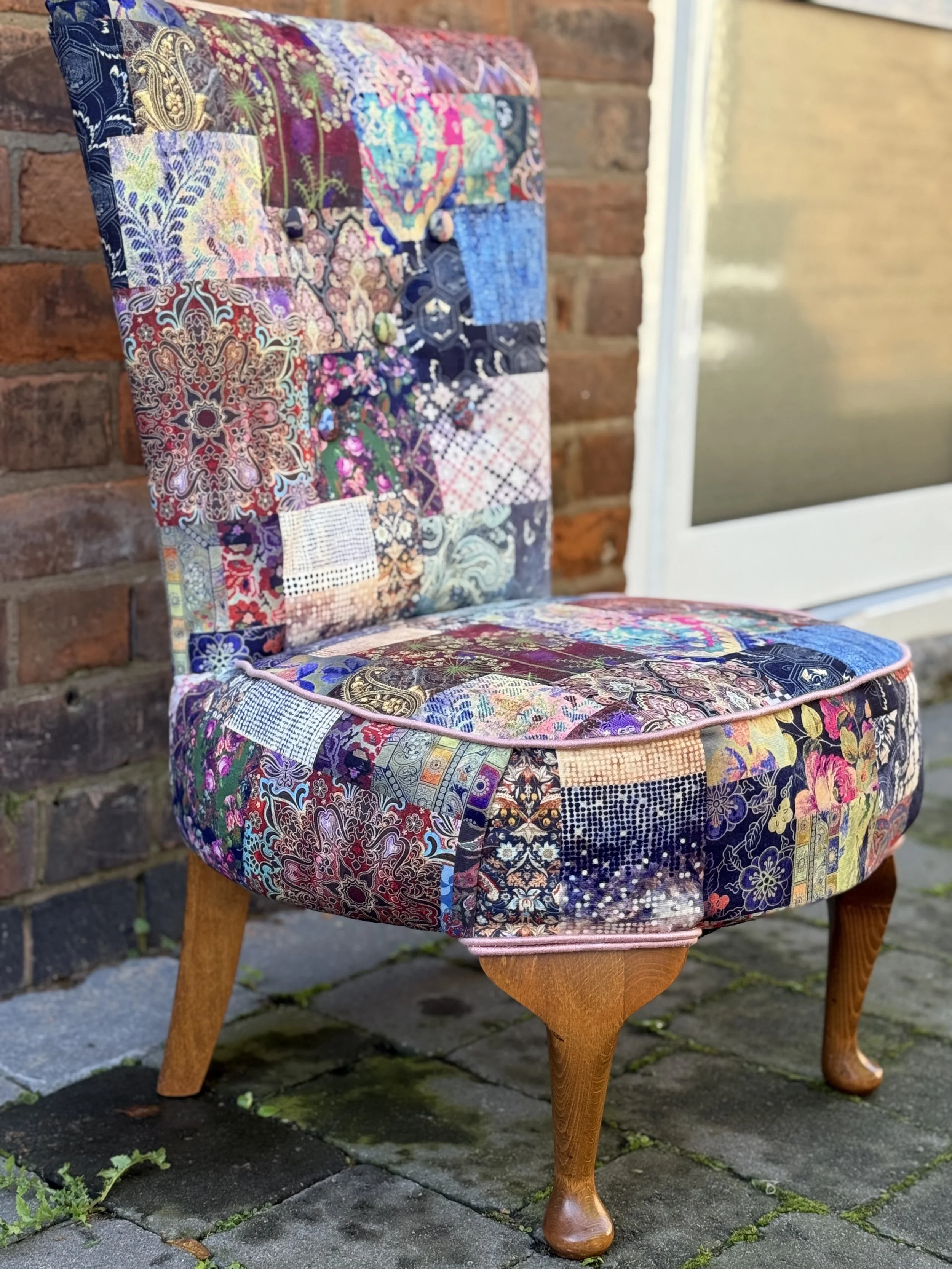 A chair with a patchwork fabric cover composed of various colorful and patterned fabric squares, placed outdoors on a stone patio against a brick wall and a window.