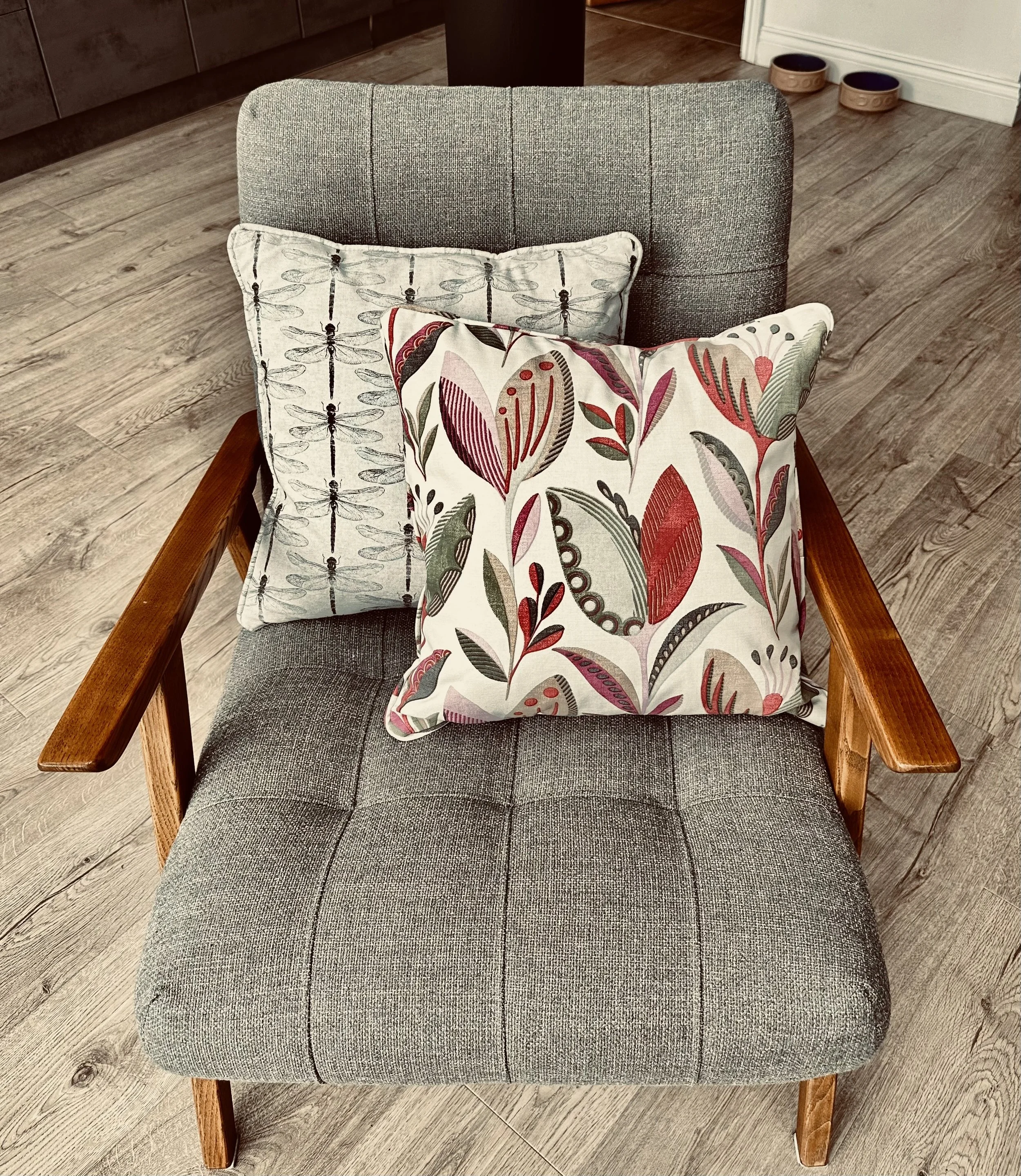 Mid-century modern gray armchair with two decorative cushions, one with a floral pattern and the other with dragonfly design, on a wooden floor.