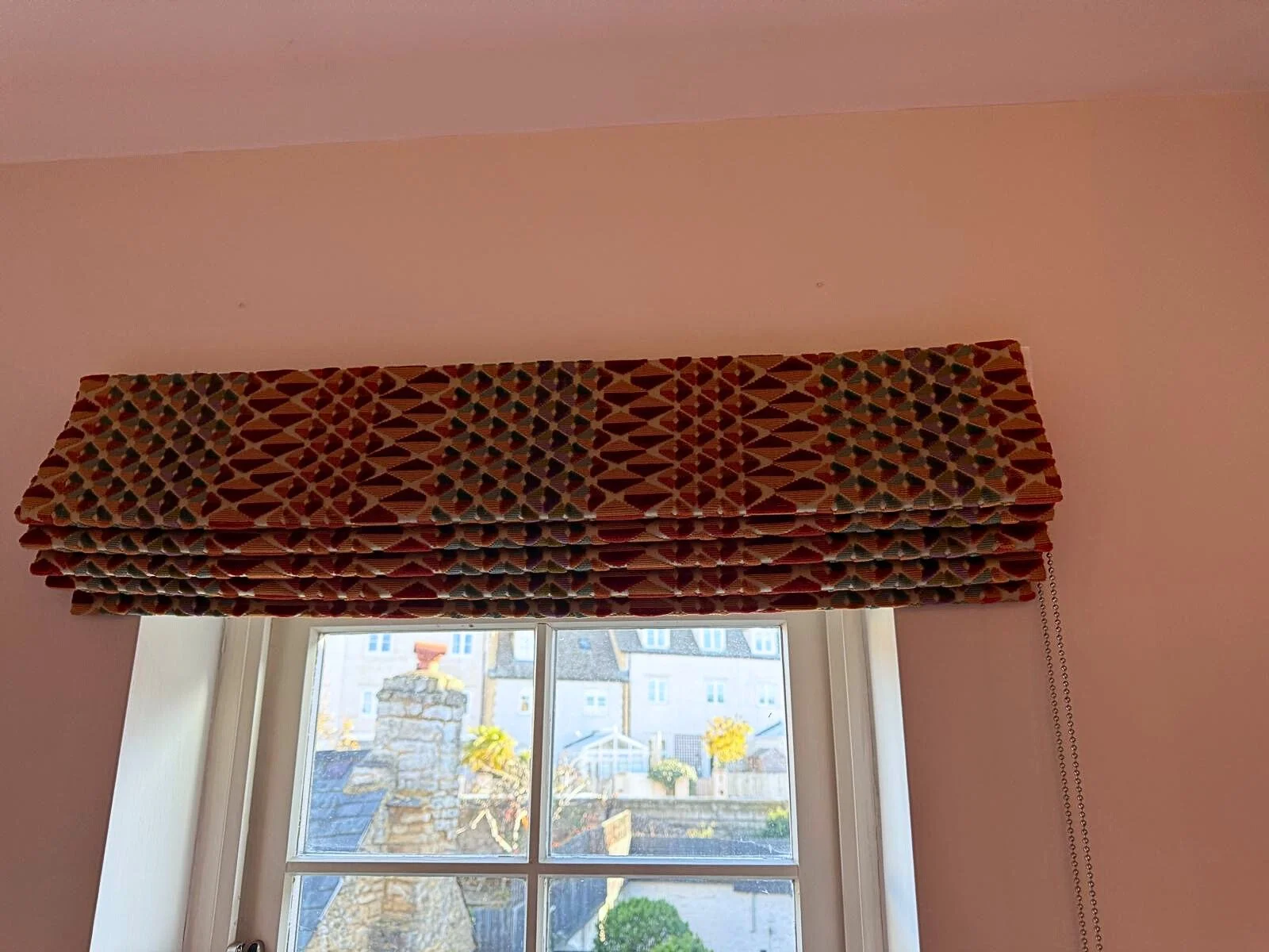 Interior view of a pink room with a window covered by a gathered patterned curtain. Outside the window, rooftops and trees are visible.