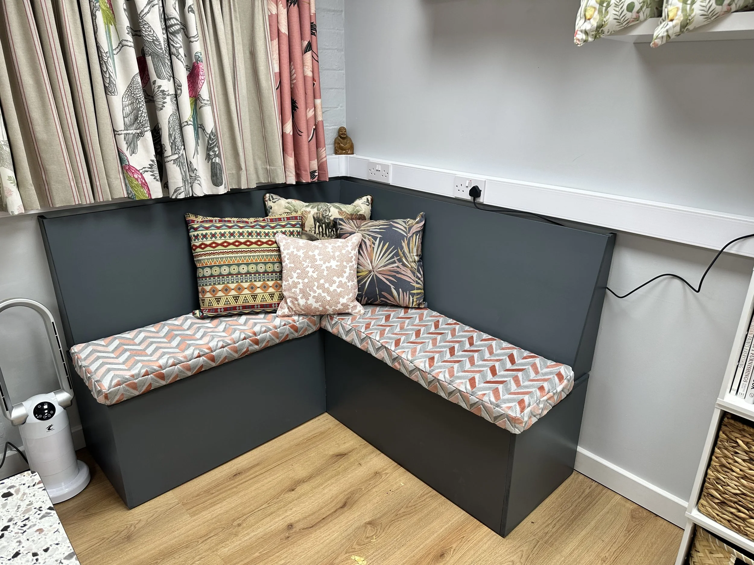 Colorful corner bench seating with patterned cushions and drapes, wooden floor, an air purifier, and a small statue on a white ledge.