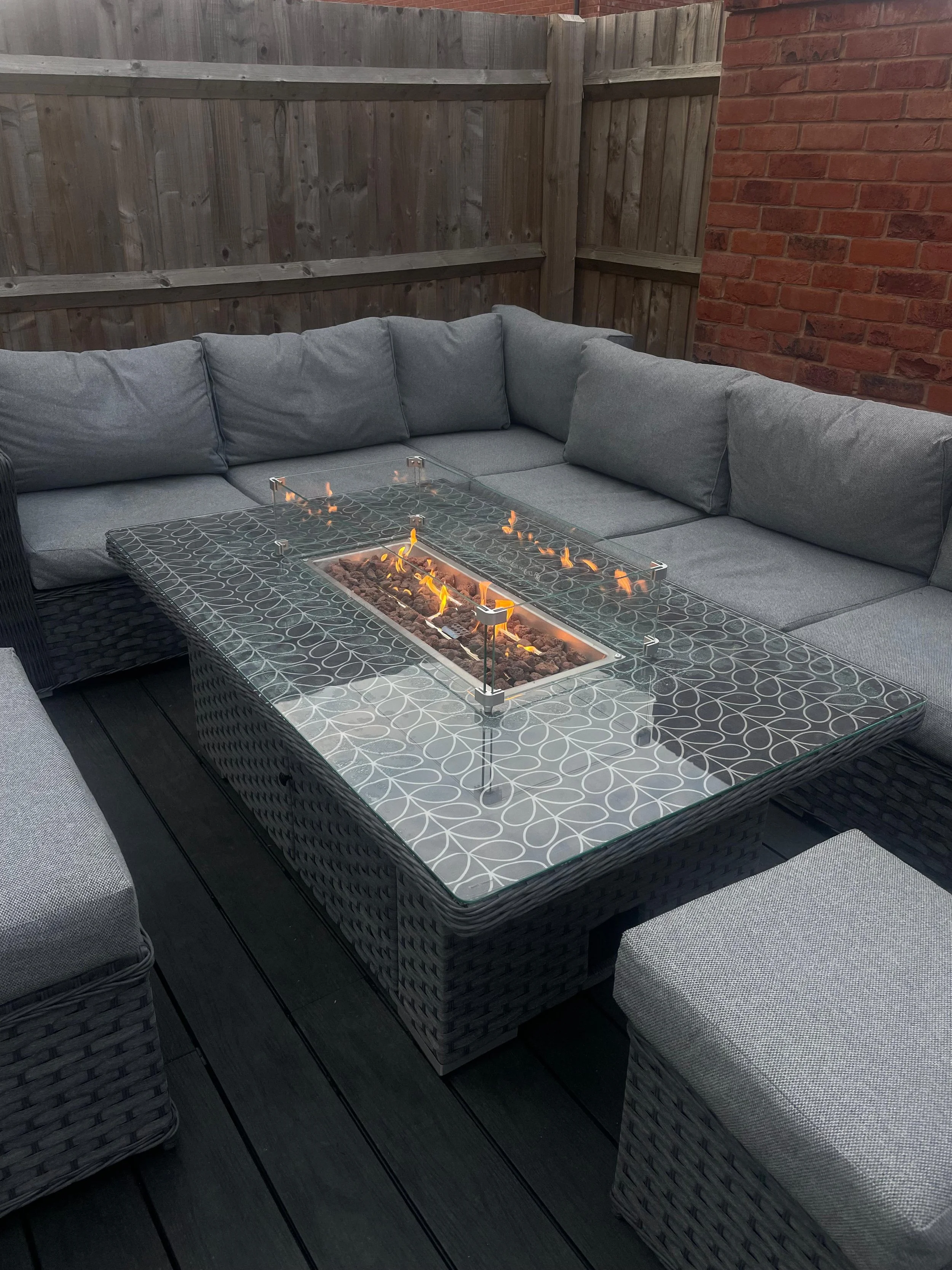 Outdoor patio with a rectangular wicker sectional sofa with gray cushions, a fire table with a glass cover, and a small stool on a dark wood deck enclosed by a wooden fence and a brick wall.