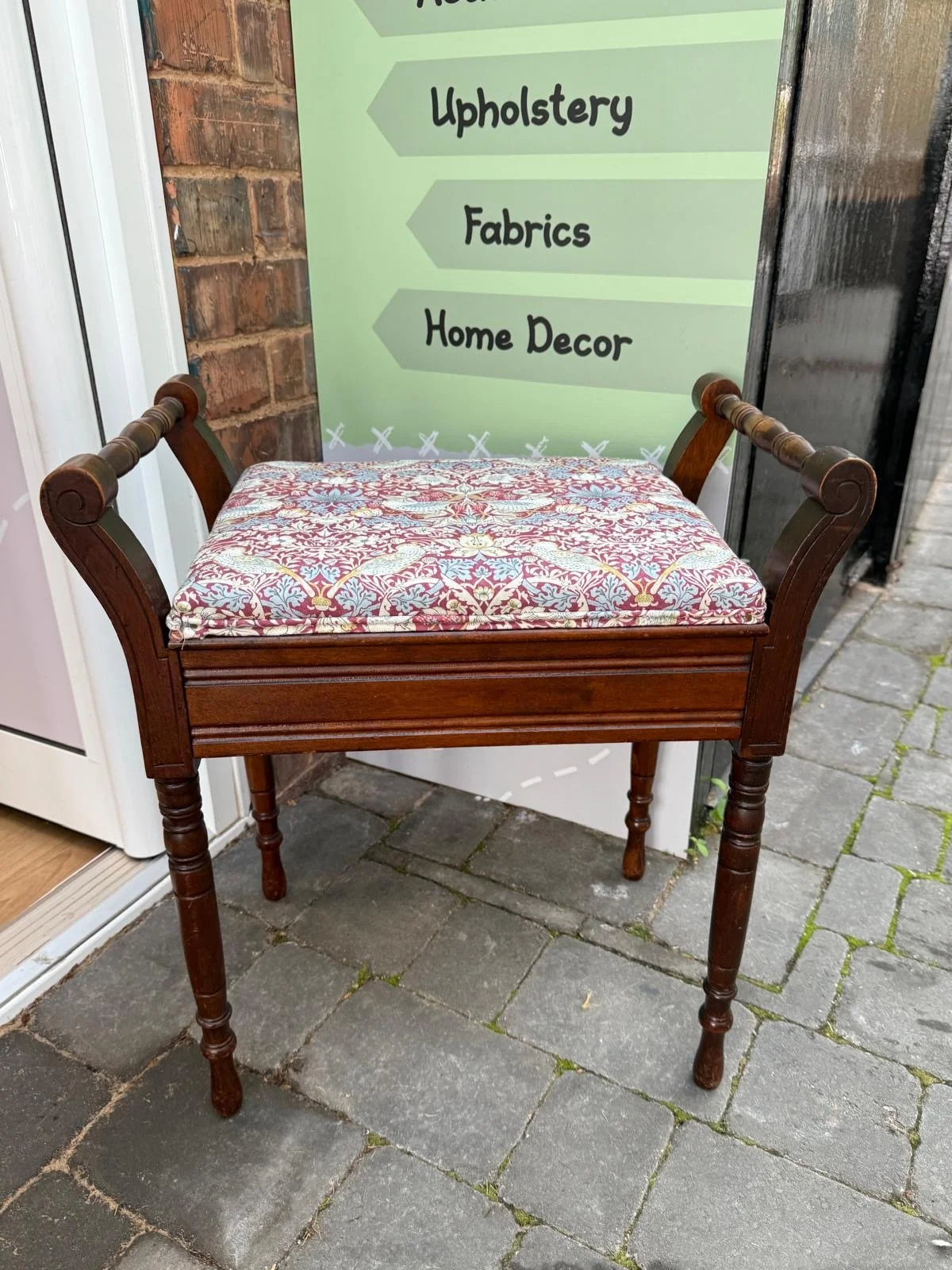 A wooden chair with a padded seat cushion featuring a red, white, and blue pattern, placed outdoors on a paved surface, with a store sign in the background listing categories such as upholstery, fabrics, and home decor.