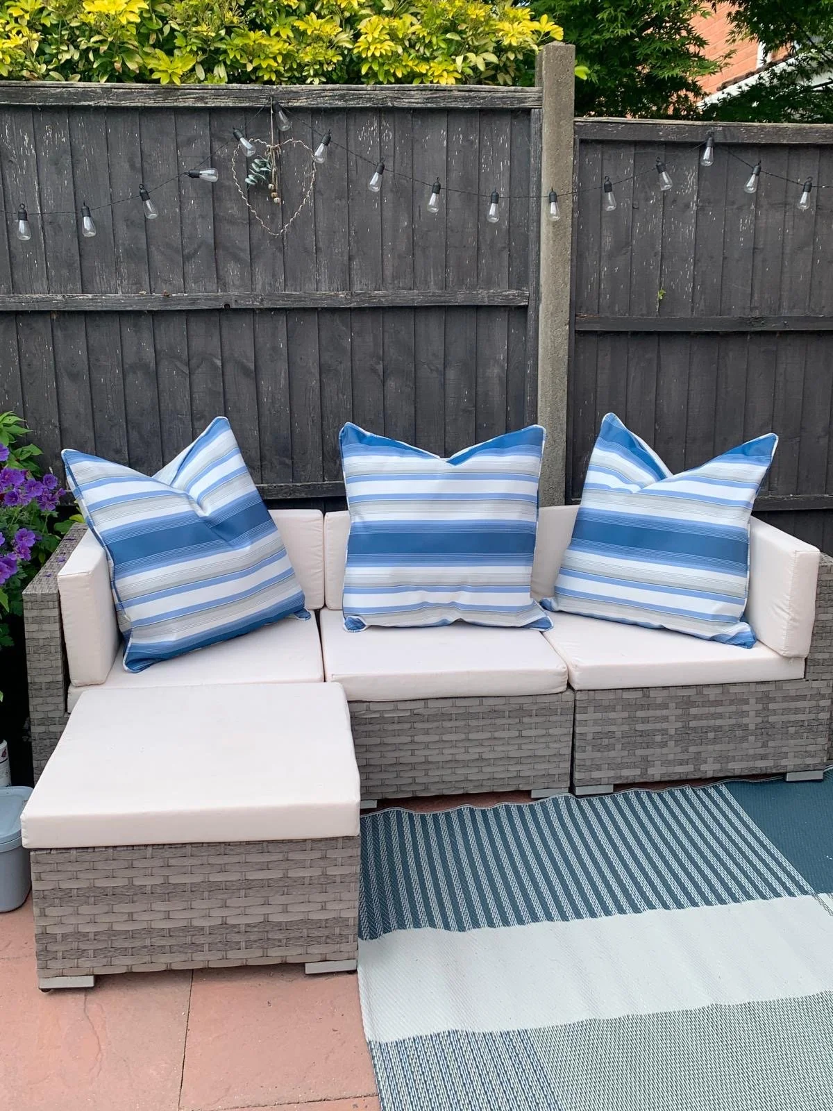 Outdoor wicker patio furniture with white cushions and blue striped pillows, set against a dark wooden fence with string lights and greenery. A striped outdoor rug lies in front of the seating area.