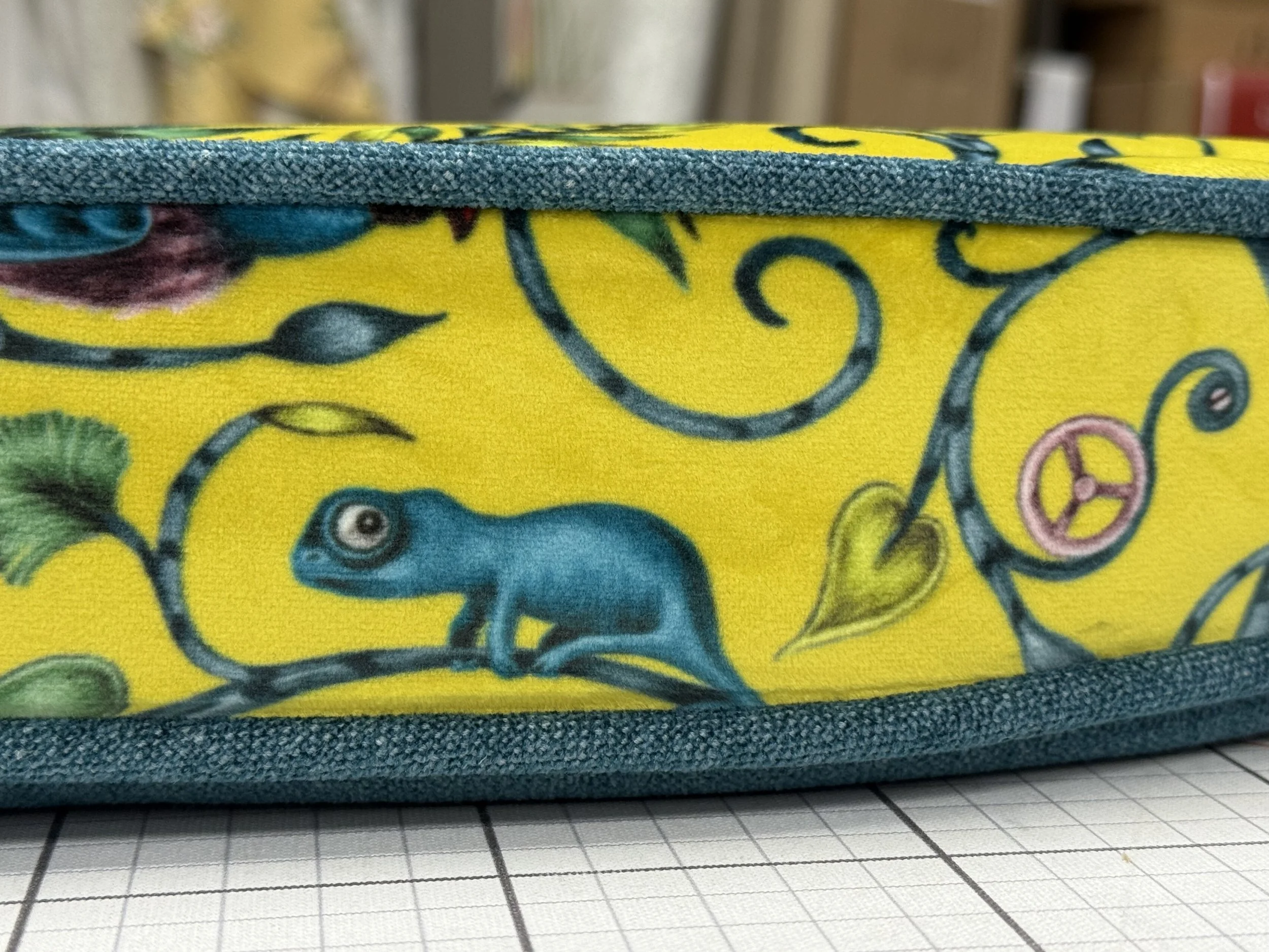 Close-up of a yellow fabric cover with a blue chameleon, green leaves, pink peace sign, and floral patterns, shown on a grid-lined surface.