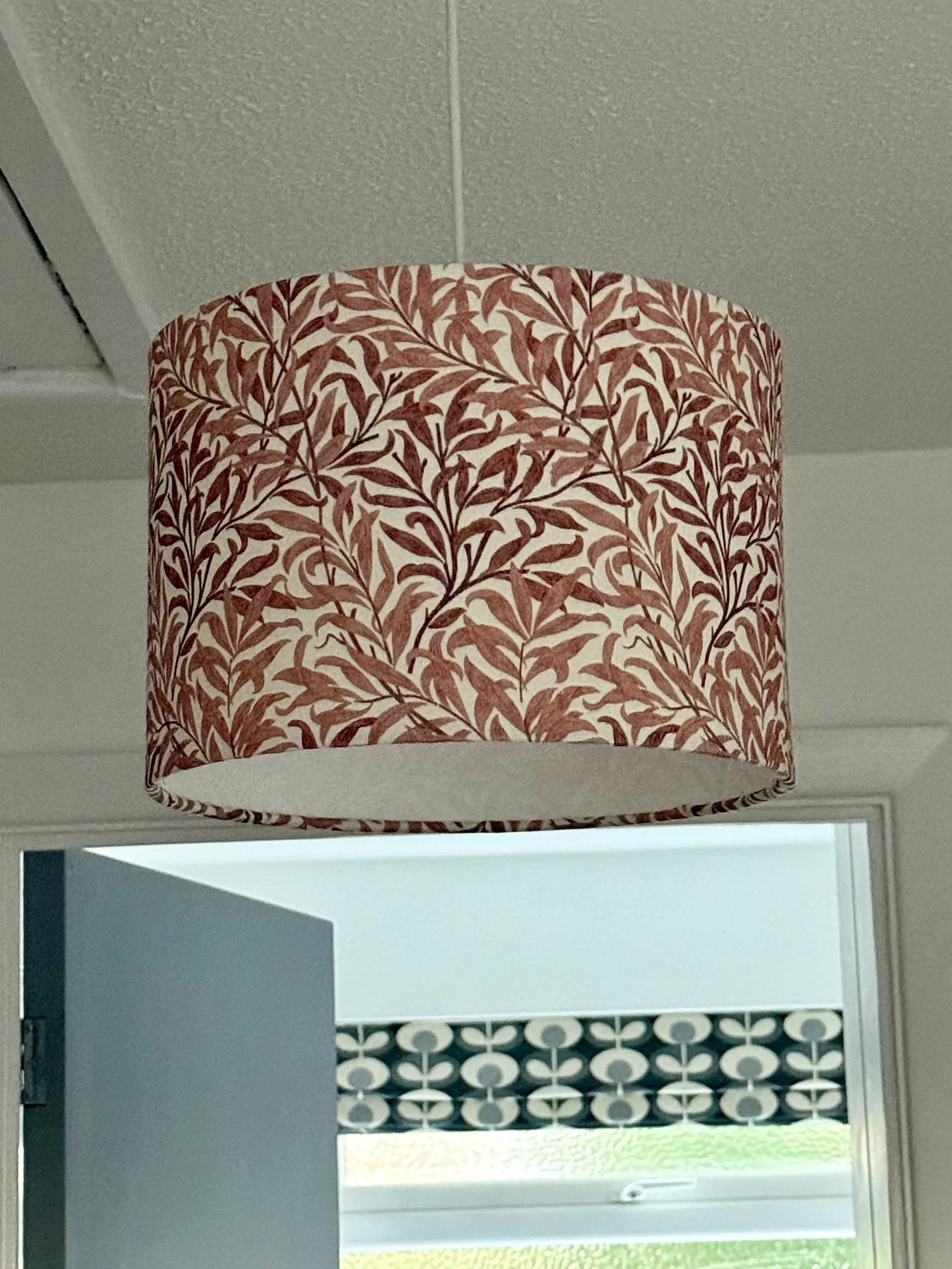 A lampshade with a red and beige leaf pattern hanging from the ceiling in a room with a partially open door and a window with a patterned valance.