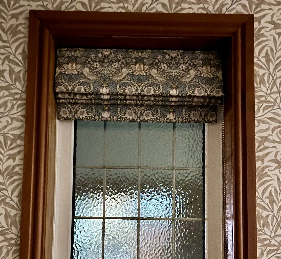 A window with frosted textured glass surrounded by a wooden frame and a fabric Roman shade with a floral and bird pattern.