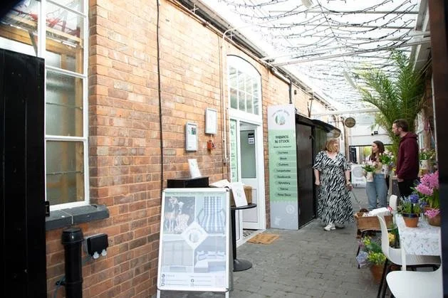 Indoor market or greenhouse with brick walls, glass ceiling, and potted plants. People are browsing or shopping, with a stand and plants on tables.