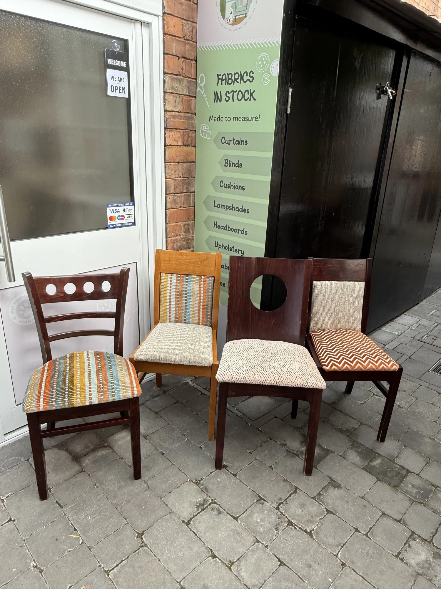 Four chairs with different colorful and patterned upholstery are arranged outside beside a brick wall and a glass door, with a green sign listing fabric types in stock.
