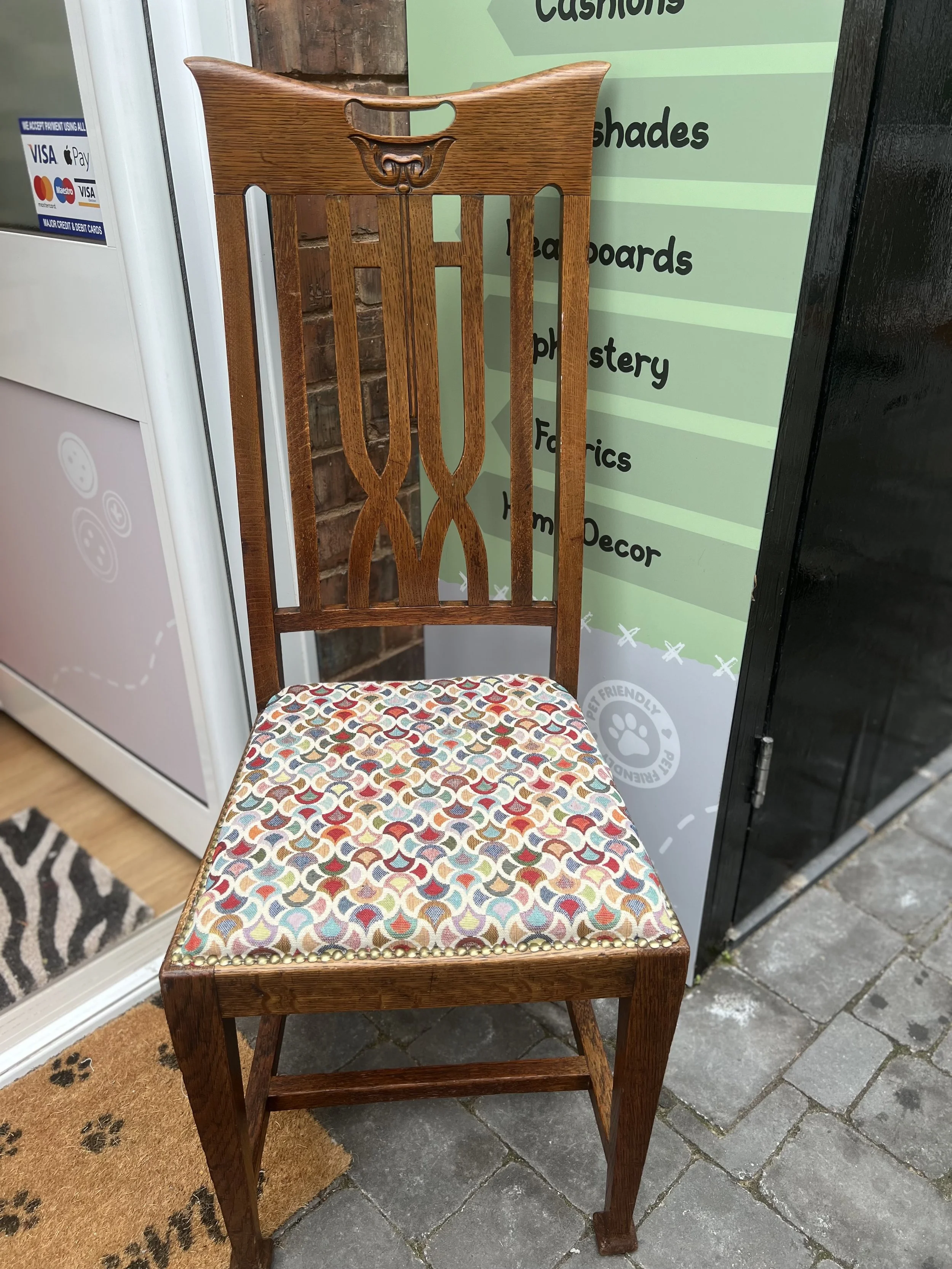 Wooden chair with a decorative backrest and a cushion with a multicolored geometric pattern, placed outside near a sign.