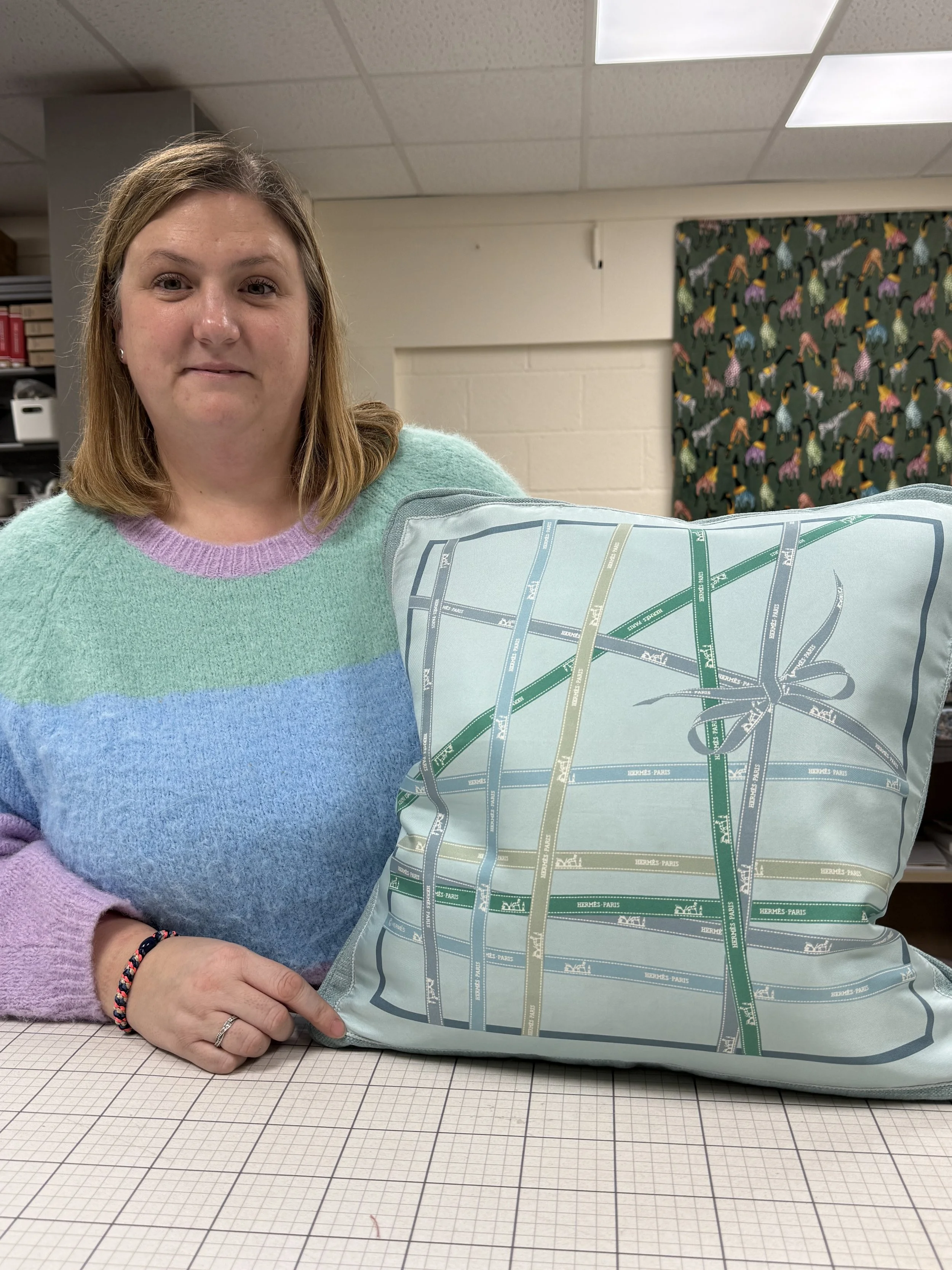 A woman sitting at a table in an indoor setting, holding a large pillow with a Hermes Paris map design. She is wearing a pastel-colored sweater with light purple, blue, and green sections, and has shoulder-length light brown hair.