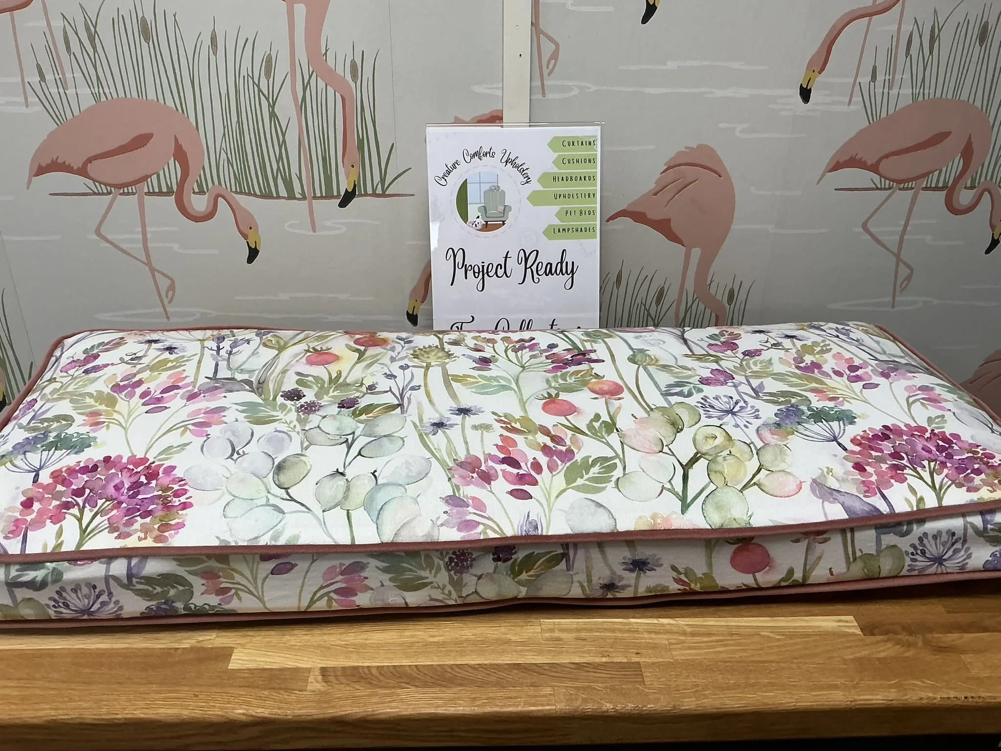 Floral-patterned mattress displayed on a wooden surface with flamingo-themed wallpaper in the background. A sign indicates the store is project ready for home furnishings.