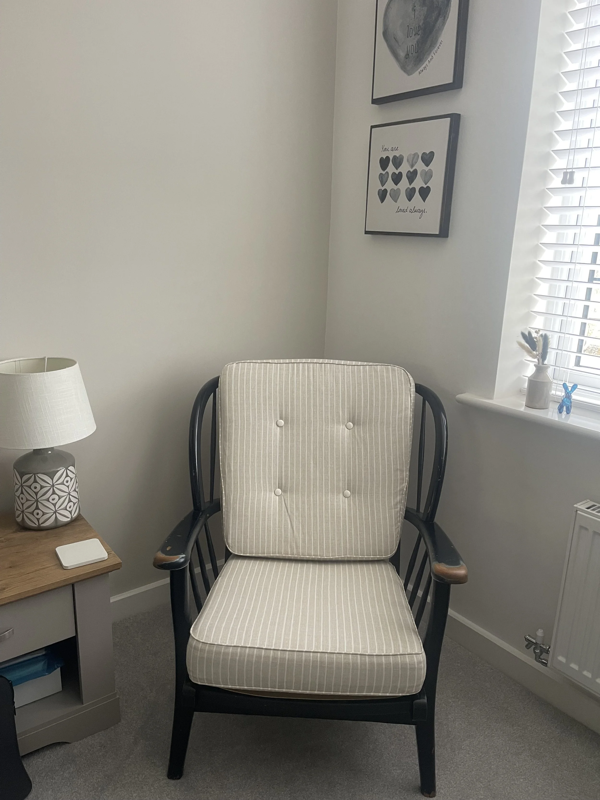 Cozy room corner with patterned lamp, wooden side table, striped cushioned chair, heart-themed wall art, white wall, and window with blinds.
