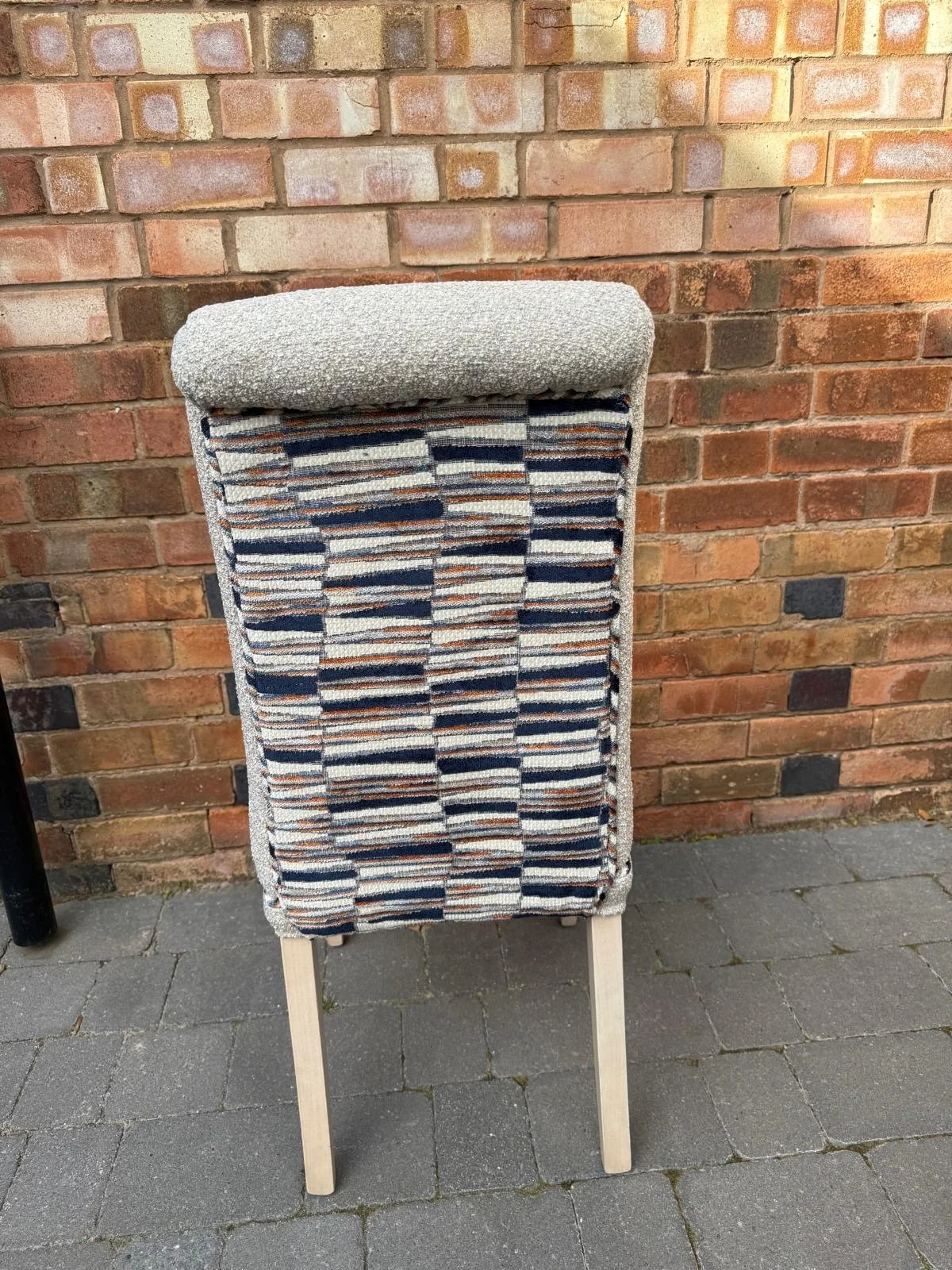A chair with a fabric backrest featuring a multicolored striped pattern, a plain cushion on top, and wooden legs, placed on a paved surface against a brick wall background.