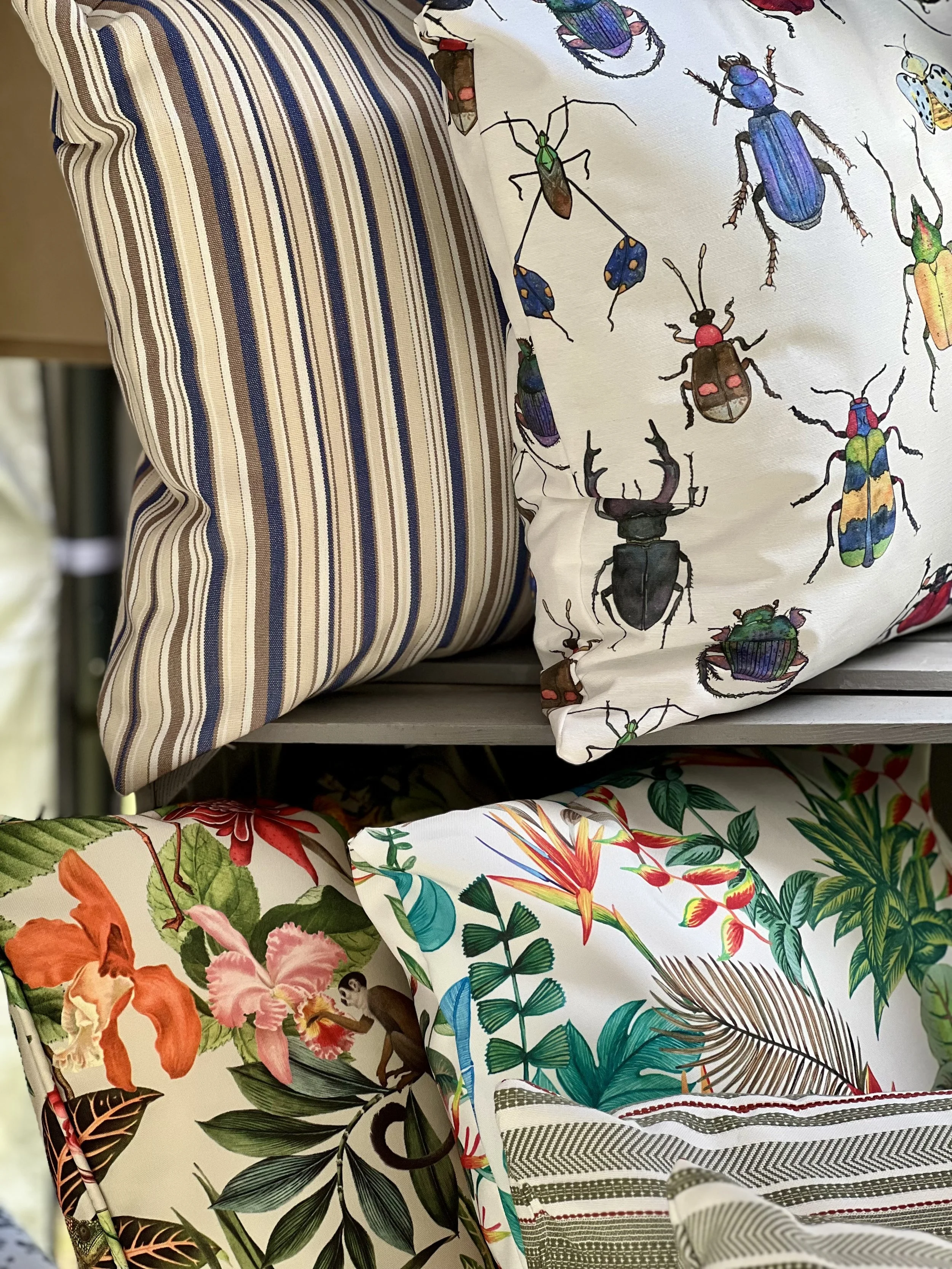 A collection of cushions with various patterns. One features stripes in shades of blue and beige, another has colorful insect illustrations, and others display tropical floral and leaf designs.