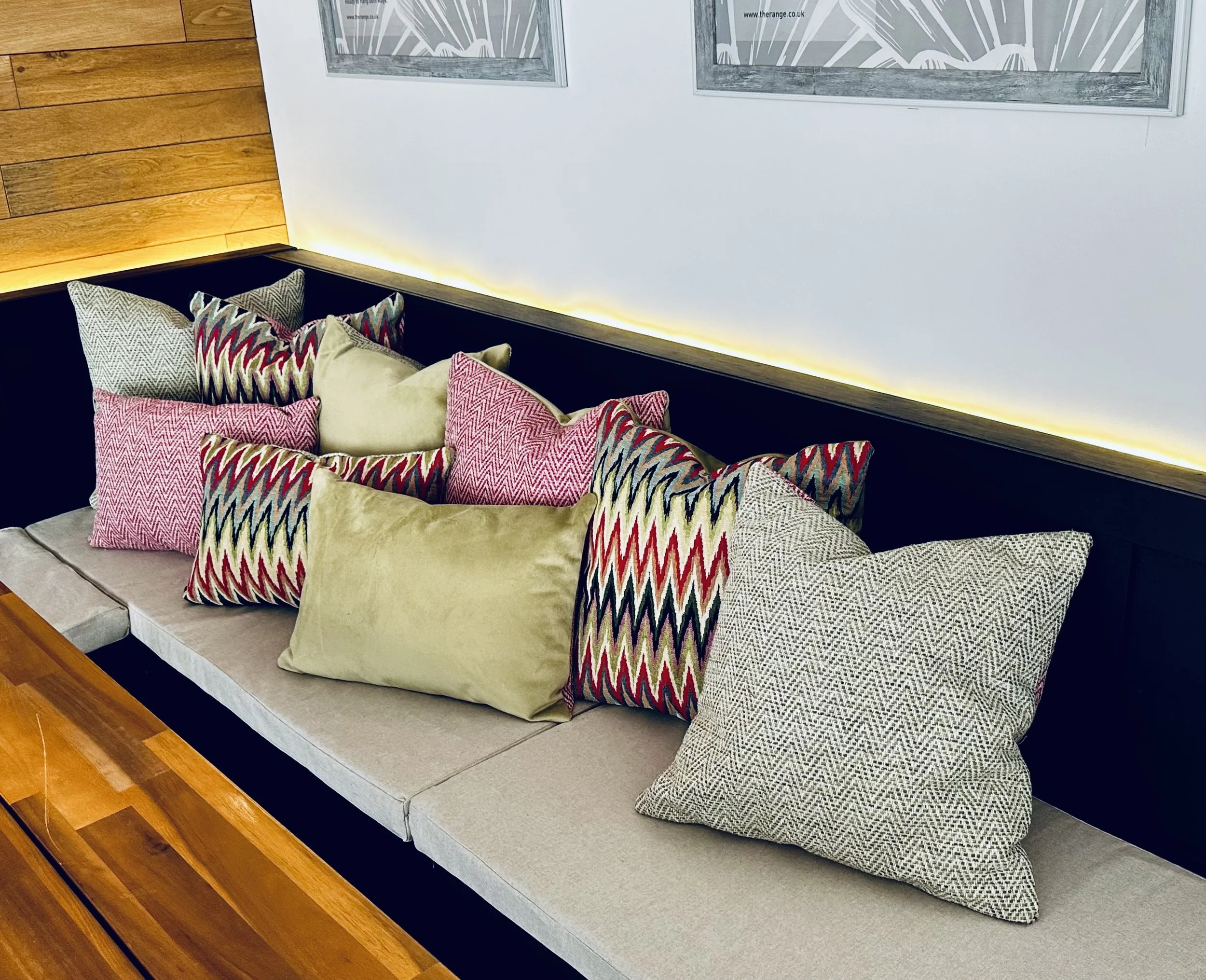 A bench with various colorful and patterned cushions against a white and wood-paneled wall. The bench seating has beige cushions and a wooden table is visible in the foreground.