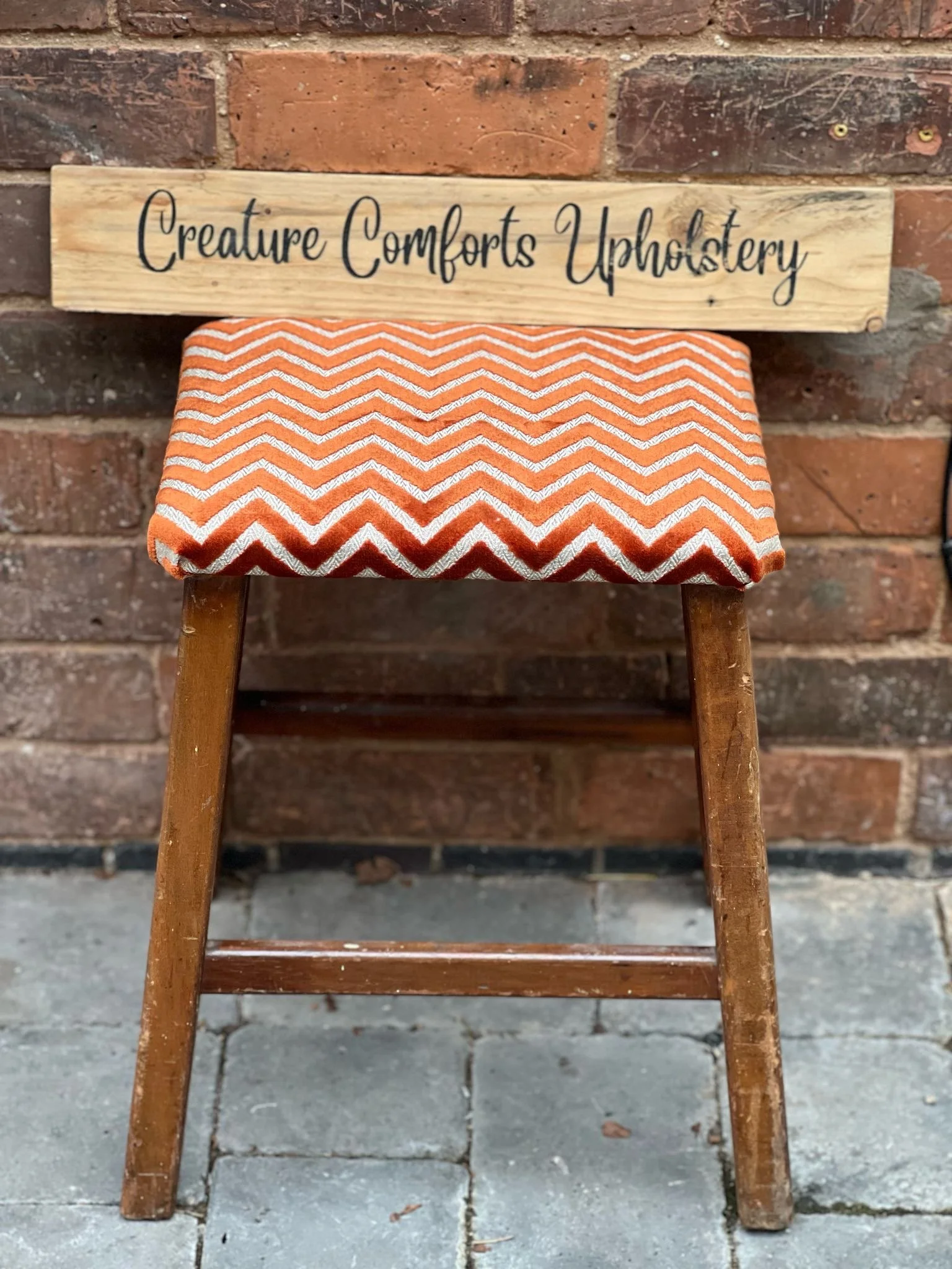 A small wooden stool with a red and white zigzag patterned cushion on top, placed on a brick patio against a brick wall. A wooden sign with the text "Creature Comforts Upholstery" is resting on the back of the stool.