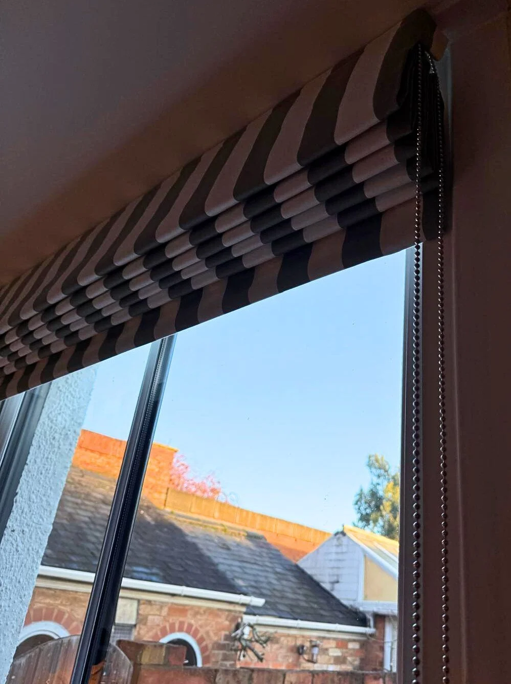 Close-up of a window with a striped fabric valance and a chain pull, showing rooftops and a blue sky outside.