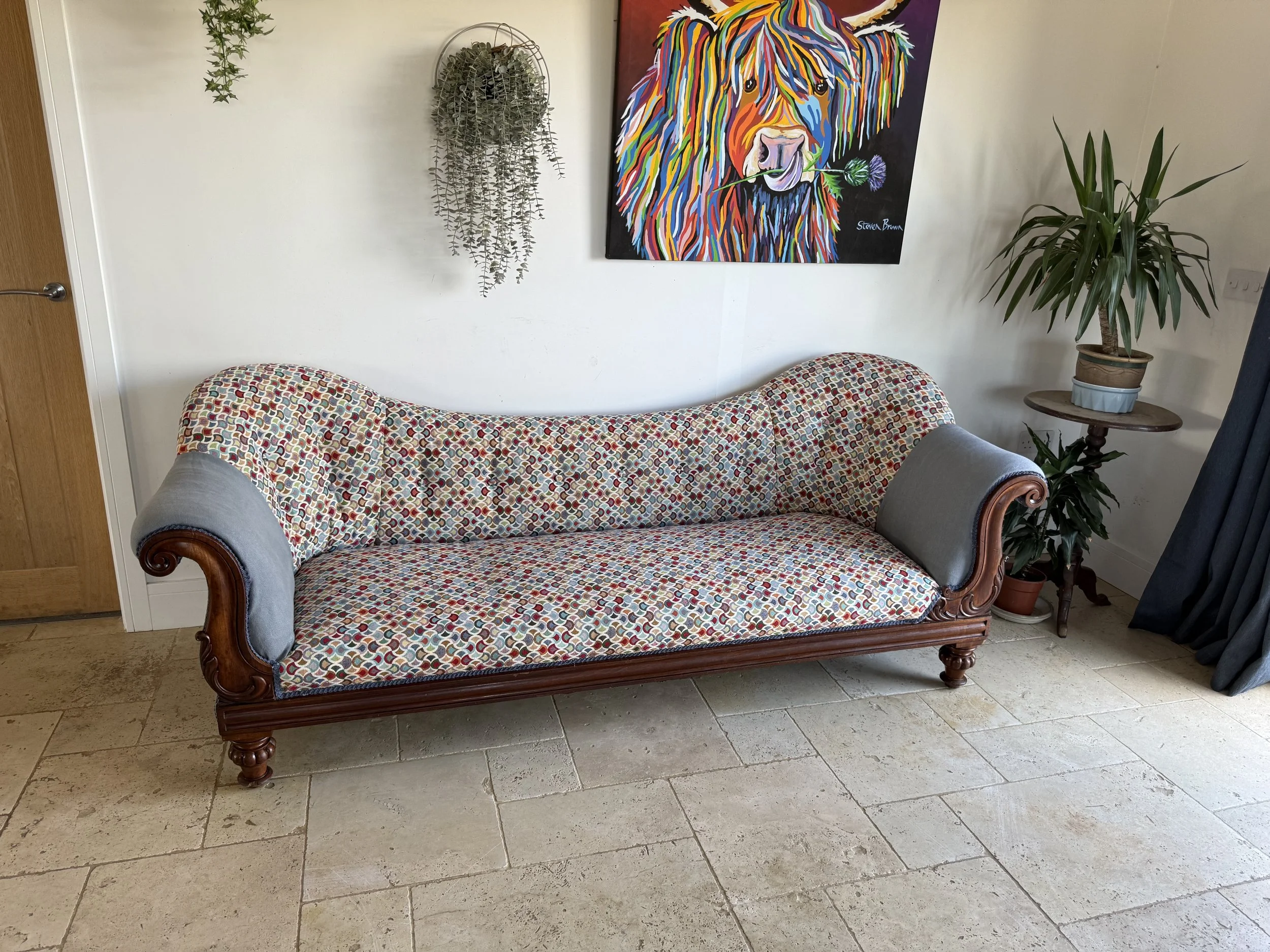 A vintage style sofa with multicolored patterned fabric and gray rolled arms sits against a white wall. Above it hangs a colorful painting of a bear with a flower in its mouth. To the right of the sofa are two stacked side tables with potted plants, 