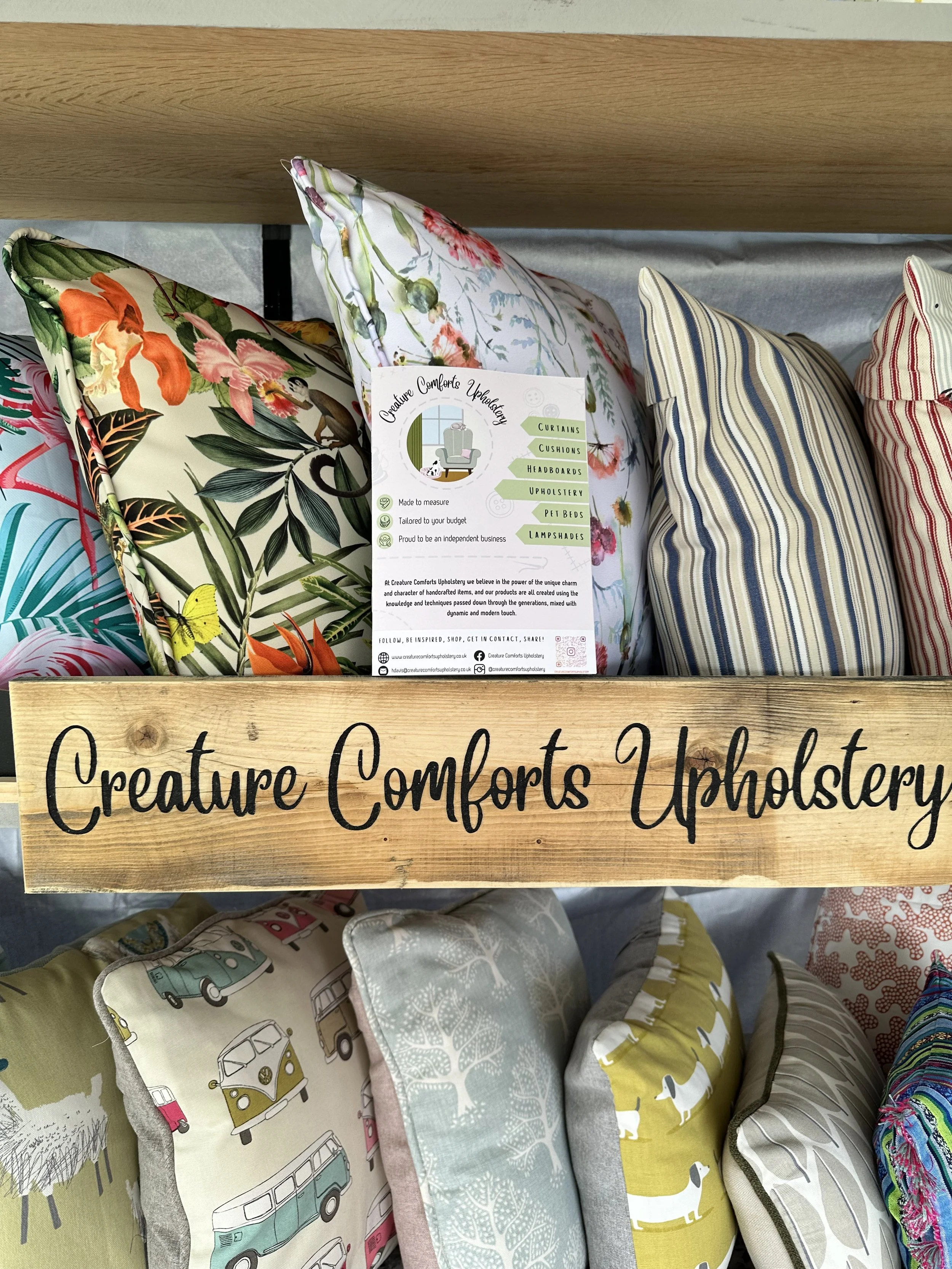 Display of various decorative pillows with different patterns and colors, under a wooden sign reading 'Creature Comforts Upholstery.' A brochure detailing services like curtains and cushions is placed among the pillows.