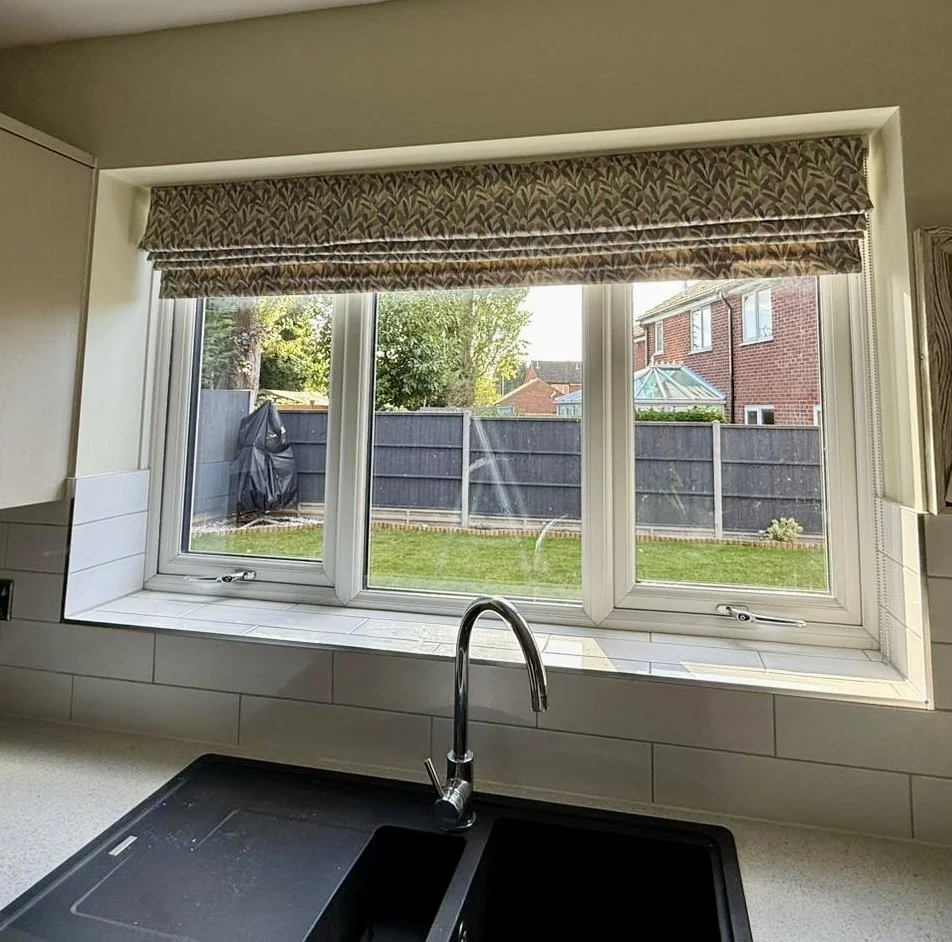Kitchen window with patterned Roman blind, view of a backyard with grass, fence, trees, and neighboring brick house.