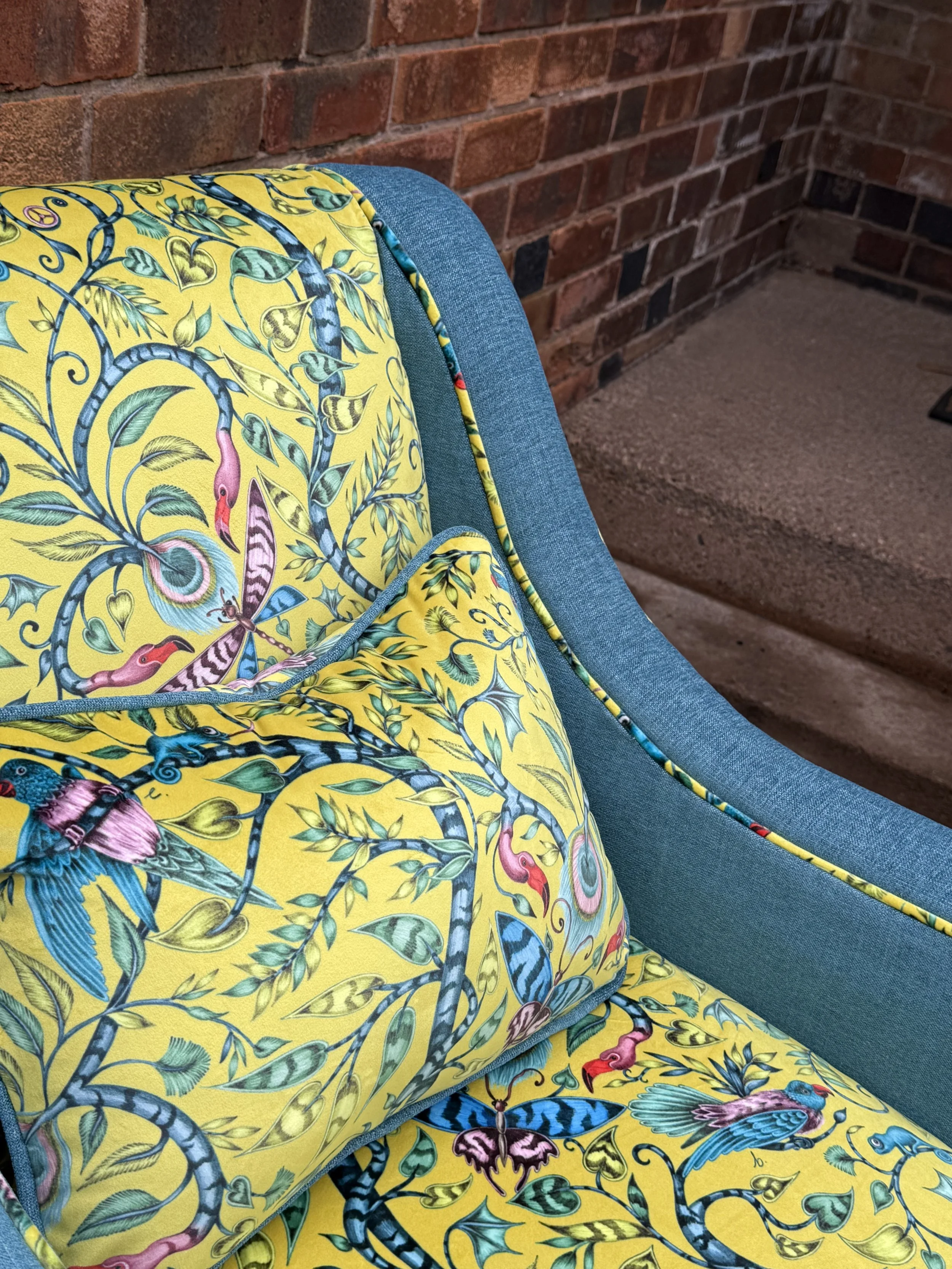 Close-up of a blue upholstered chair with a yellow fabric cover featuring a colorful, intricate bird and foliage pattern, next to a brick wall and steps.