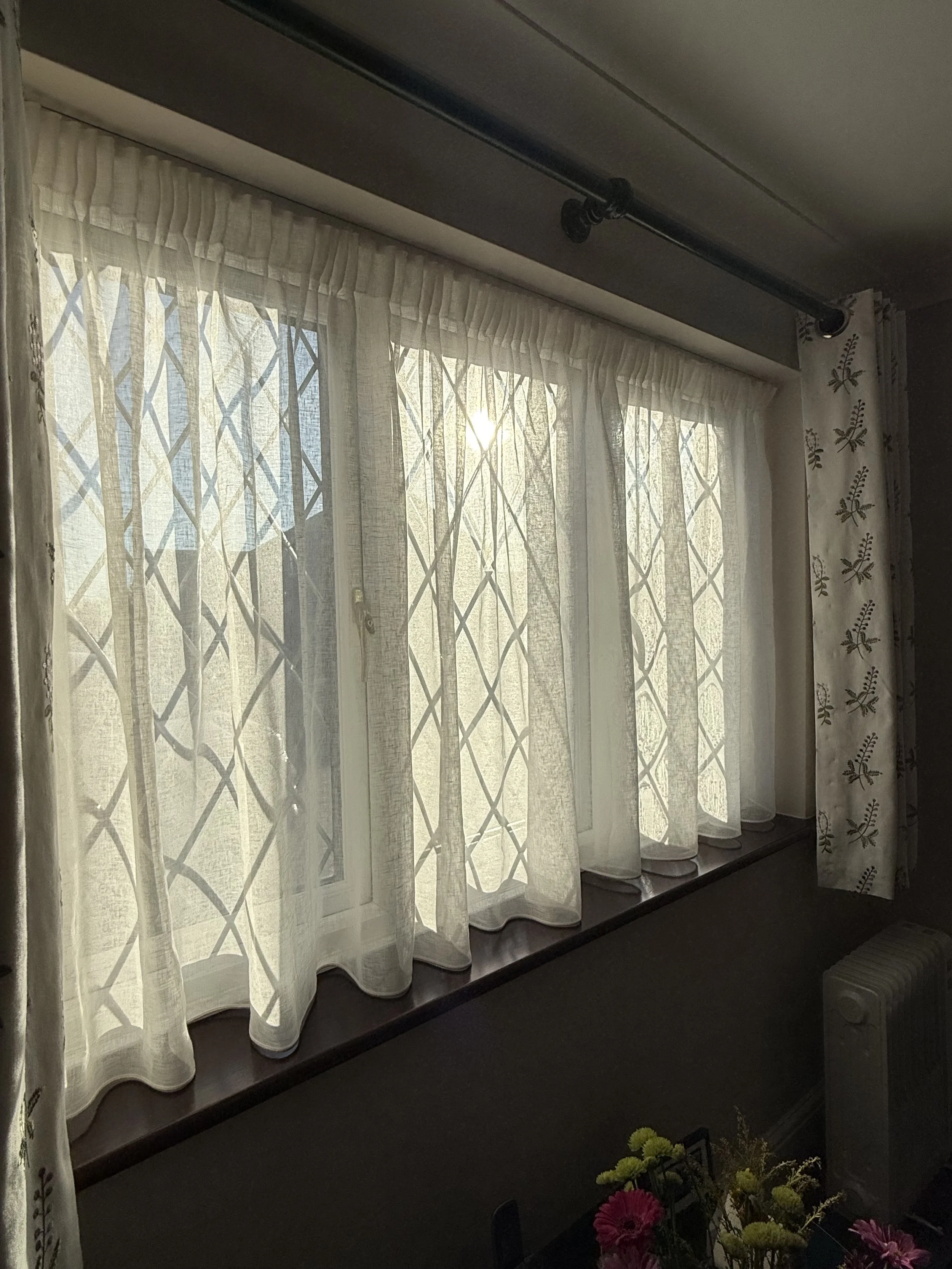 Sunlight coming through a window with white sheer curtains and decorative lattice patterned security bars outside, with floral curtains on the side and a radiator beneath the window.