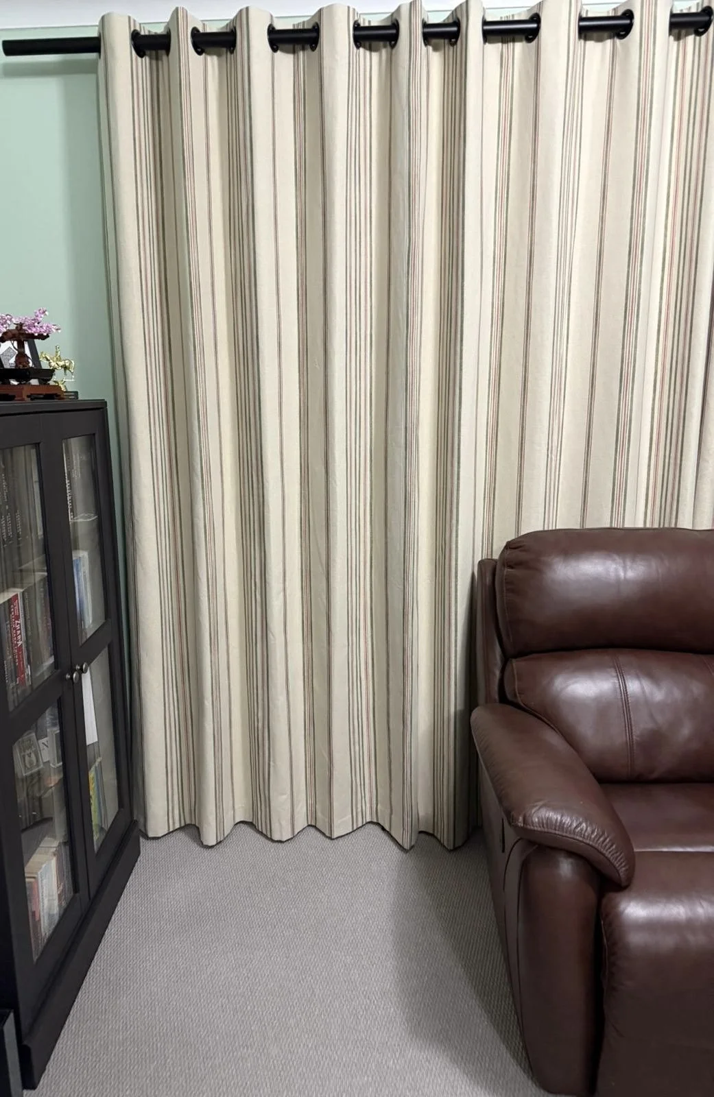 A beige curtain with vertical stripes hanging from a black curtain rod in a living room, partially covering a wall behind a brown leather armchair and a black bookshelf with glass doors.