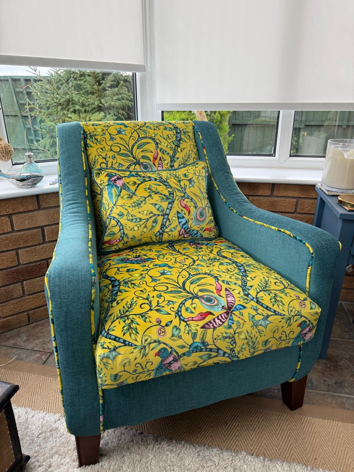 A teal armchair with floral and bird patterned upholstery, including a matching pillow, located near a window with a view of greenery outside.