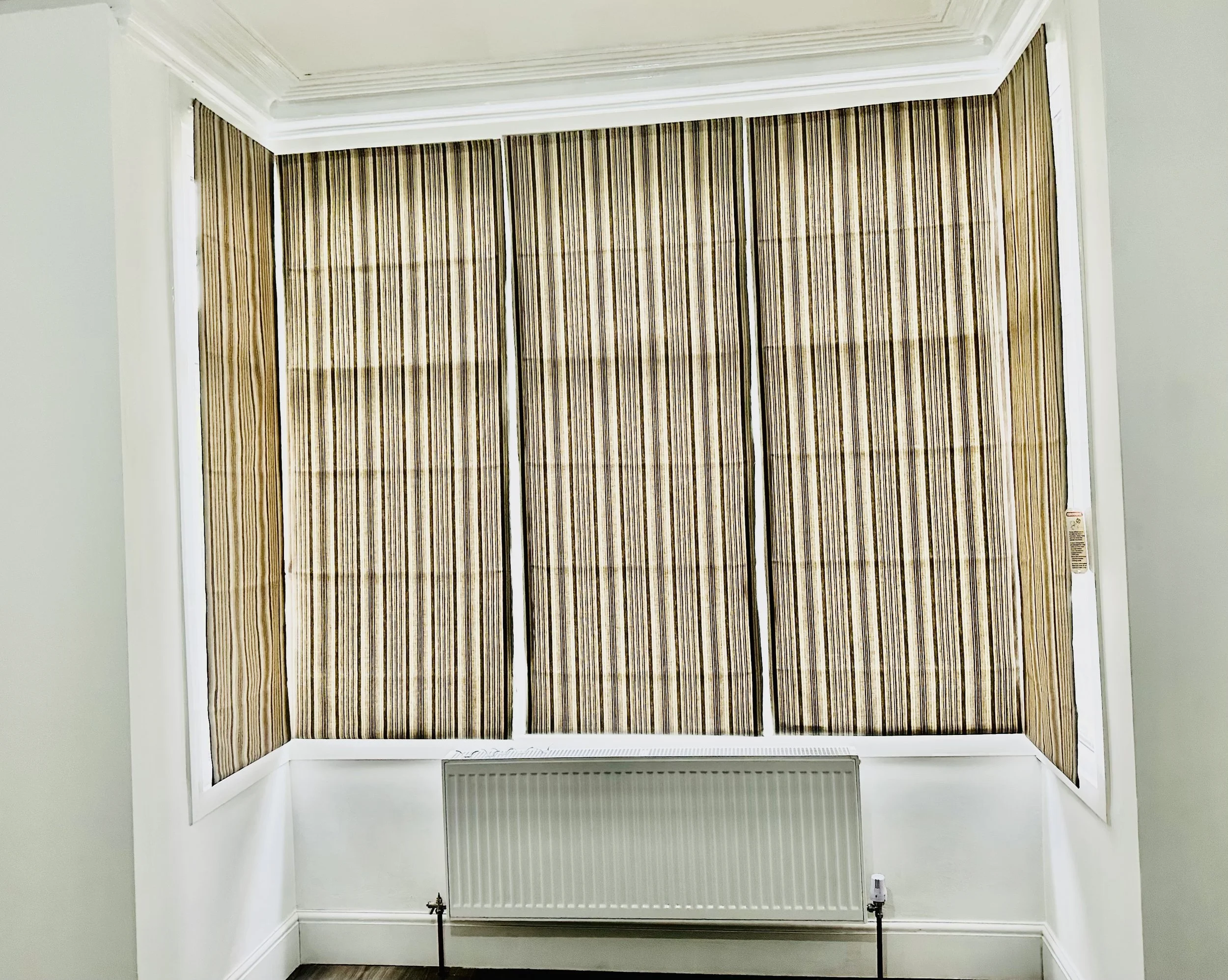 Room with striped brown window blinds covering three large windows, white walls, crown molding, and radiator below the windows.